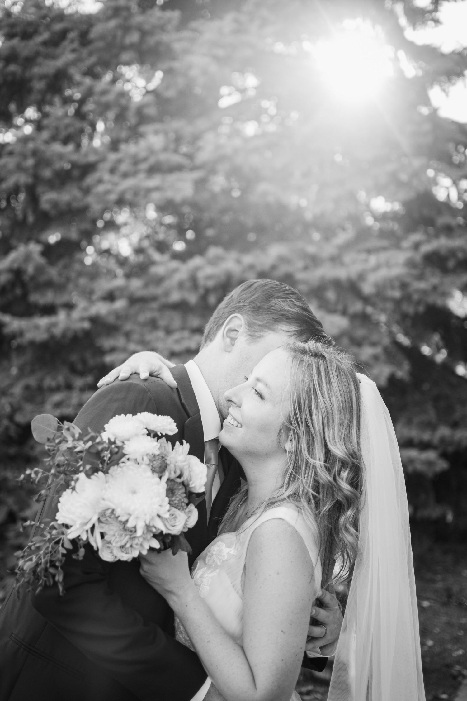 Black and white photo of a smiling bride holding bouquet while groom kisses her cheek outdoors.