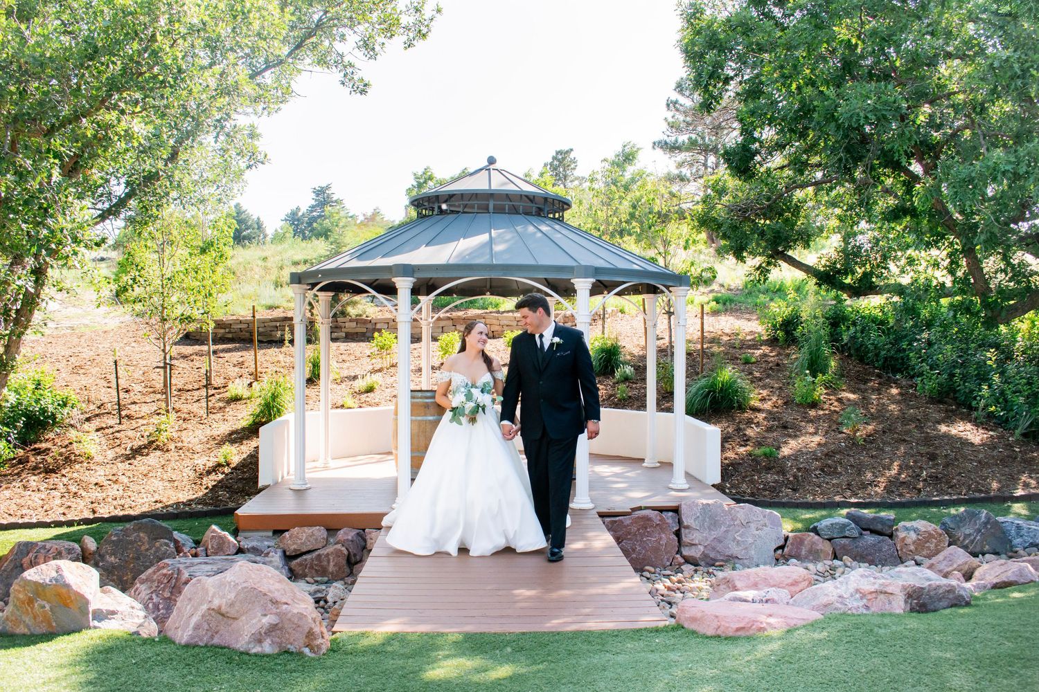 Bride and groom walking hand in hand near a white gazebo an outdoor wedding venue at Creekside Terrace by Wedgewood.