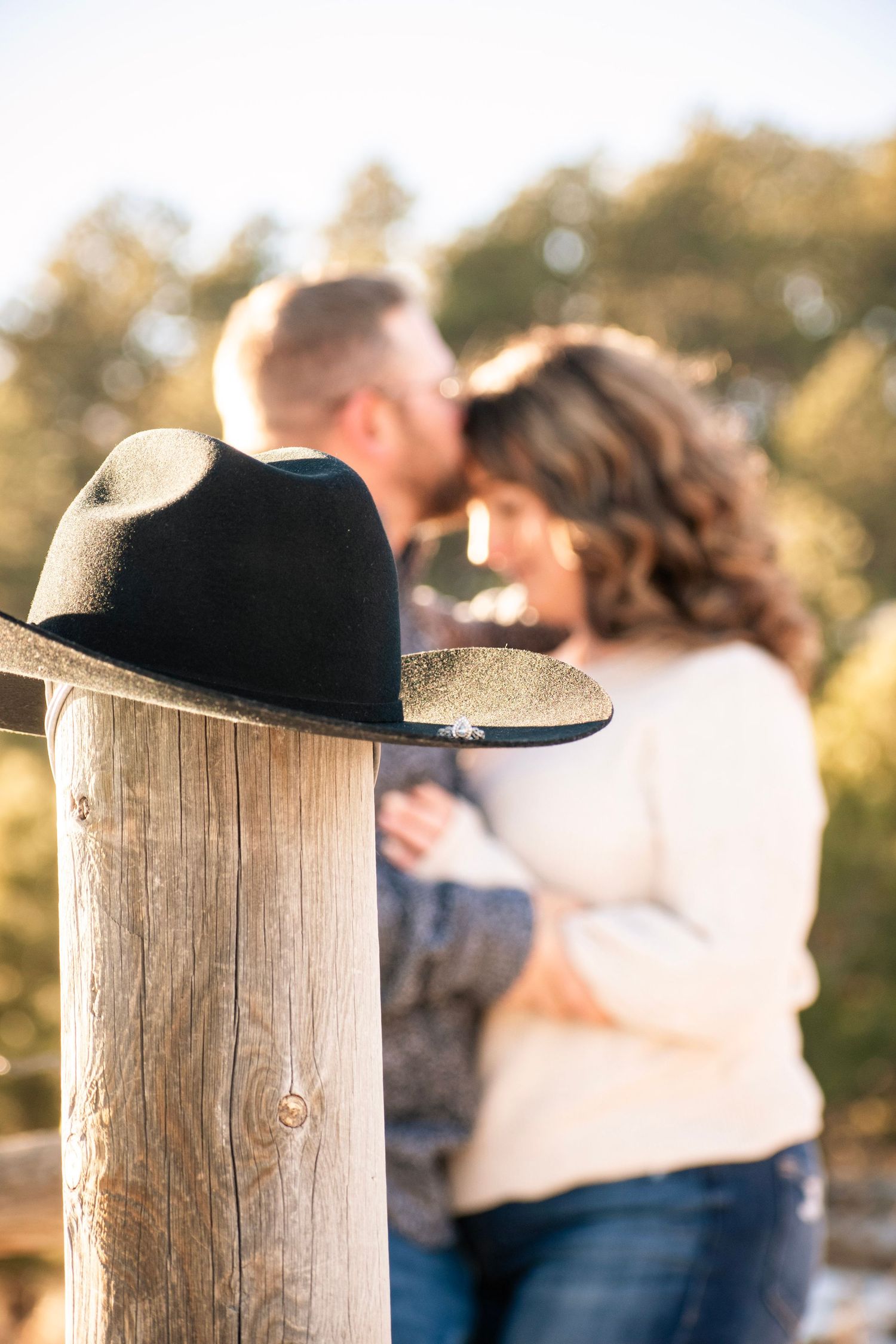 A black cowboy hat rests on a wooden post as a couple embraces and shares a kiss in a sunny outdoor setting.