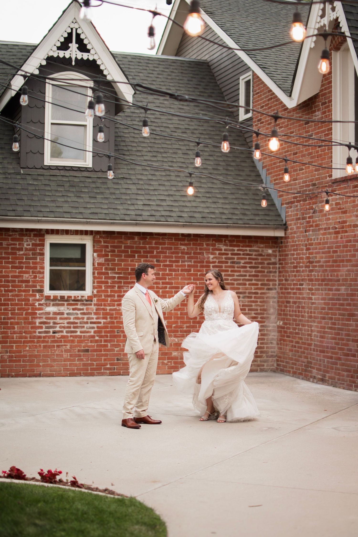 A bride and groom share a romantic dance outdoors on a patio, surrounded by string lights against a brick building.