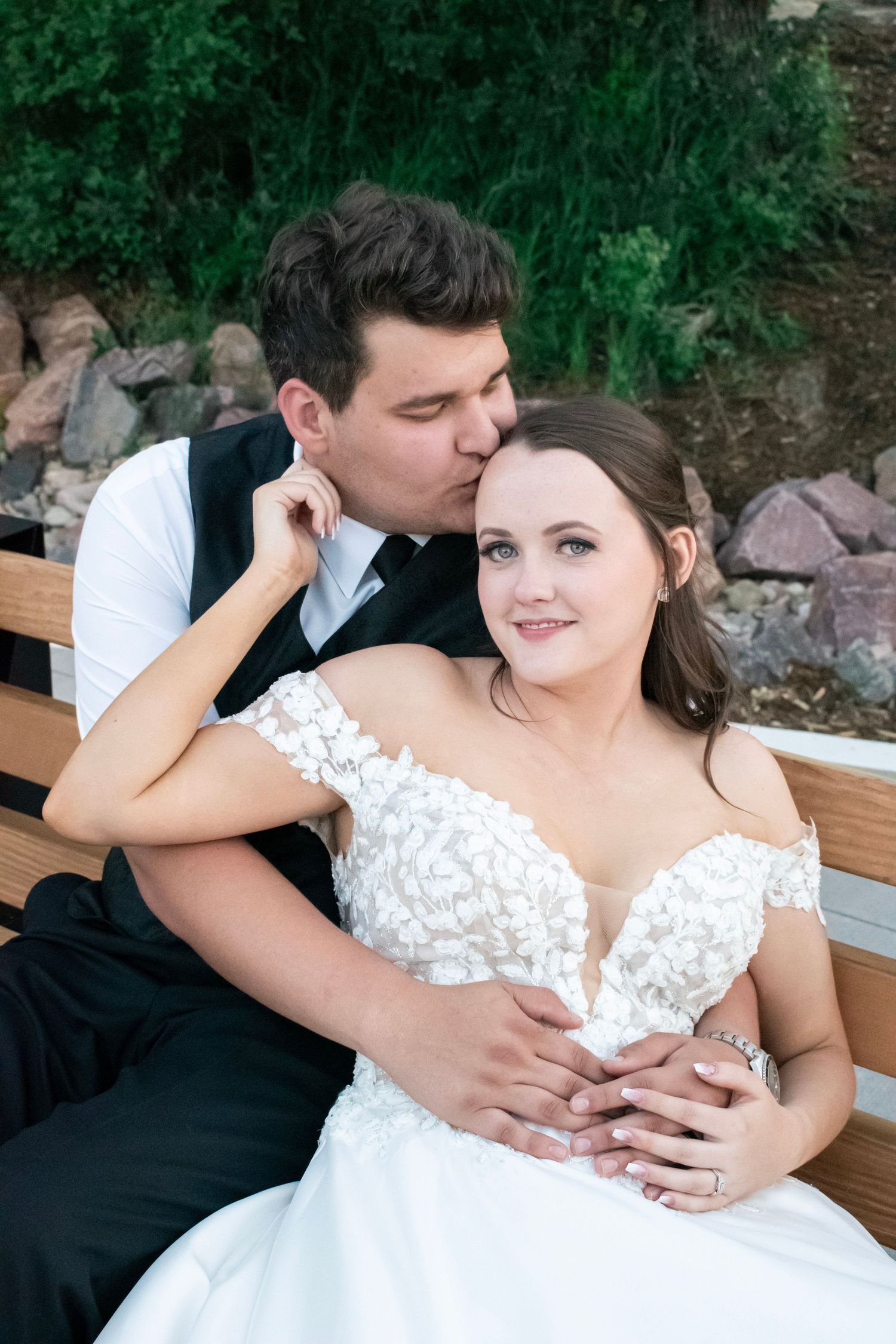 Groom kisses bride on cheek as she smiles on bench, wearing white off-shoulder lace wedding gown outdoors.