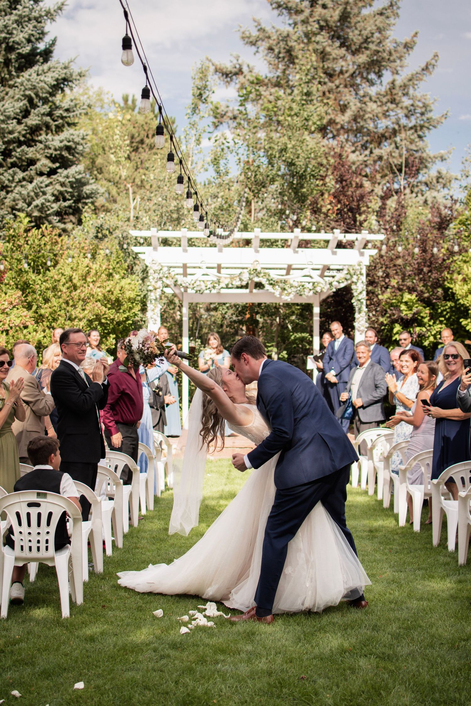Groom dips bride for a kiss as wedding guests cheer along the aisle at outdoor ceremony.