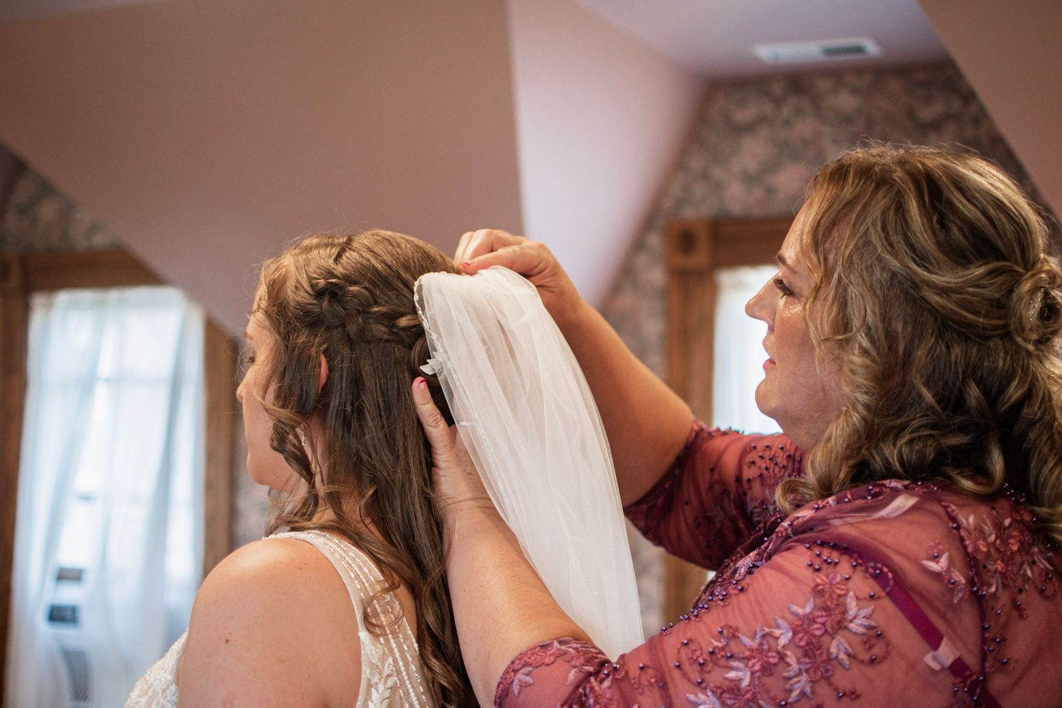 Mother carefully placing bridal veil into bride's braided updo hairstyle before wedding ceremony.