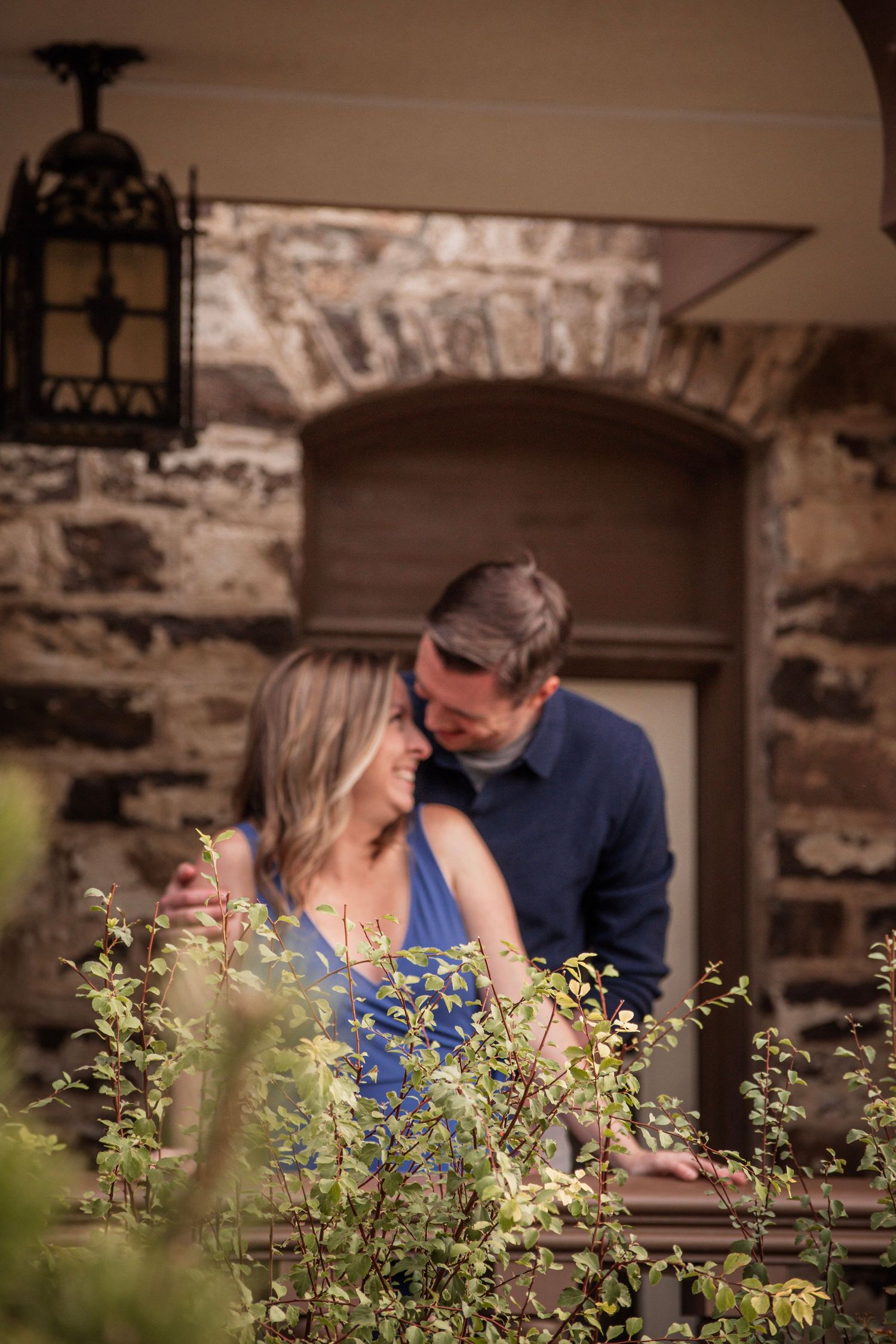 Romantic couple sharing a tender moment among lush greenery before a rustic stone building with arched doorway.