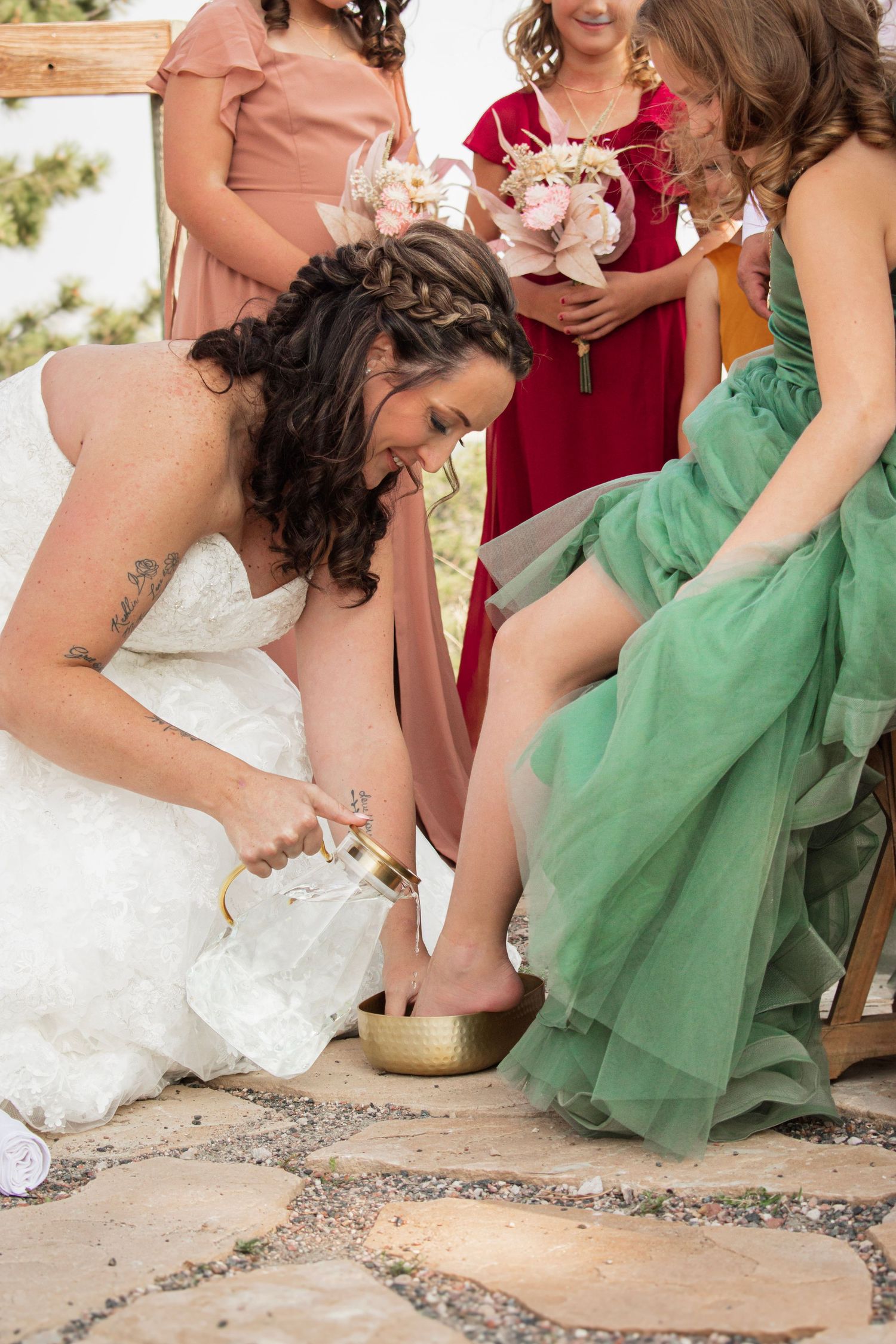 Bride performing a foot washing ceremony for bridesmaid in green dress during outdoor wedding celebration.