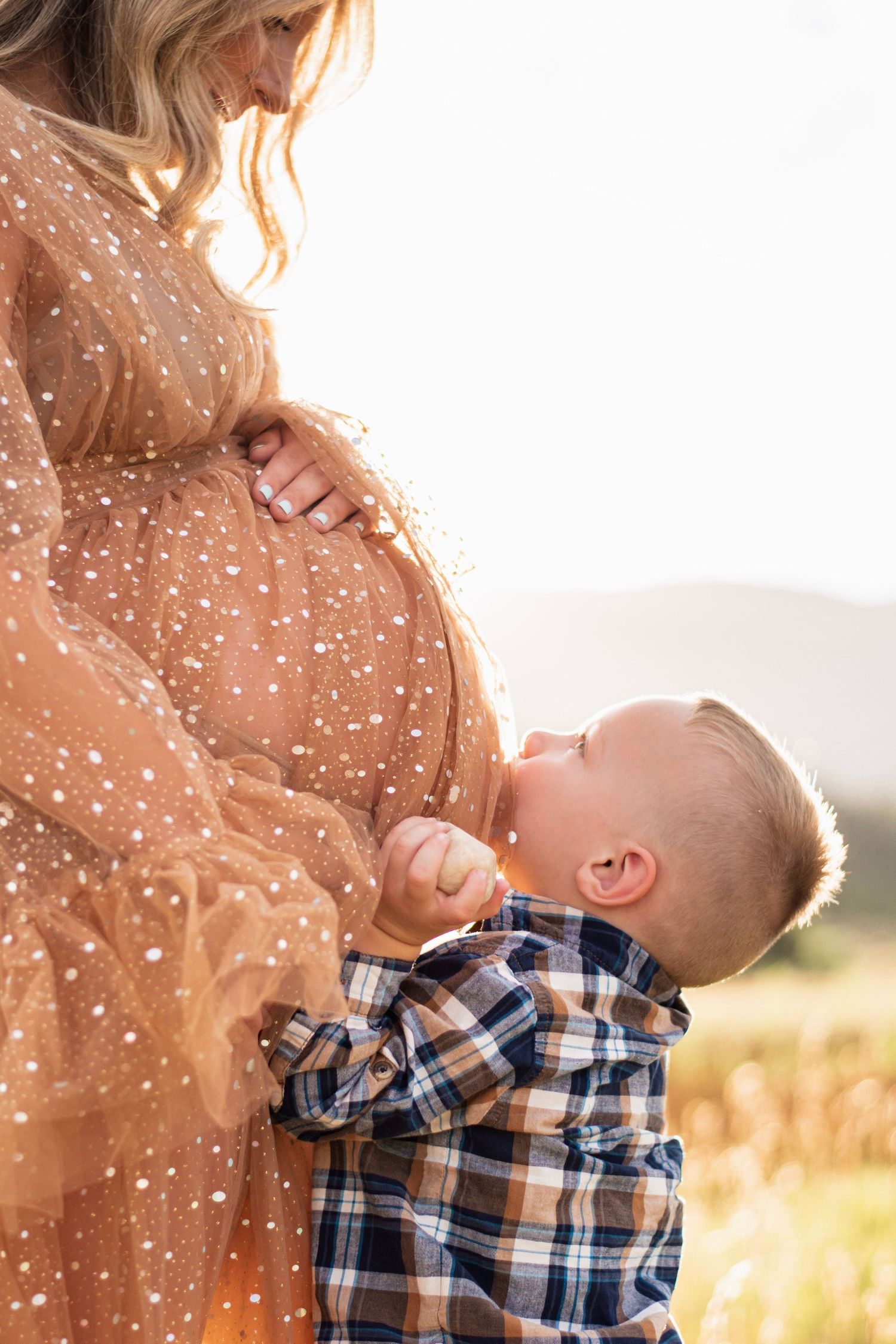 A young boy kisses a pregnant woman's belly outdoors in golden sunlight, both wearing warm-toned clothing.