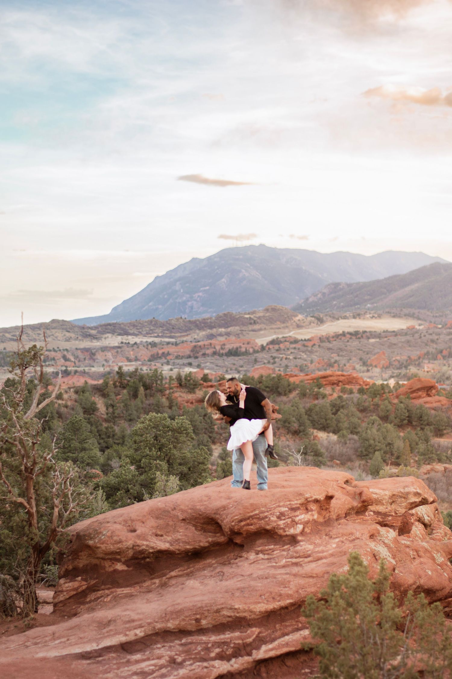 Couple sharing a romantic dip kiss on red rocks at Garden of the Gods with mountain backdrop at sunset.