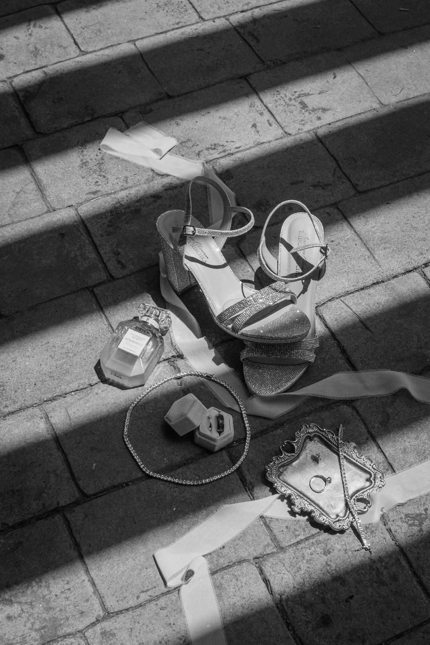 Black and white flat lay of bridal accessories including heels, perfume, ring box, and jewelry on stone steps.