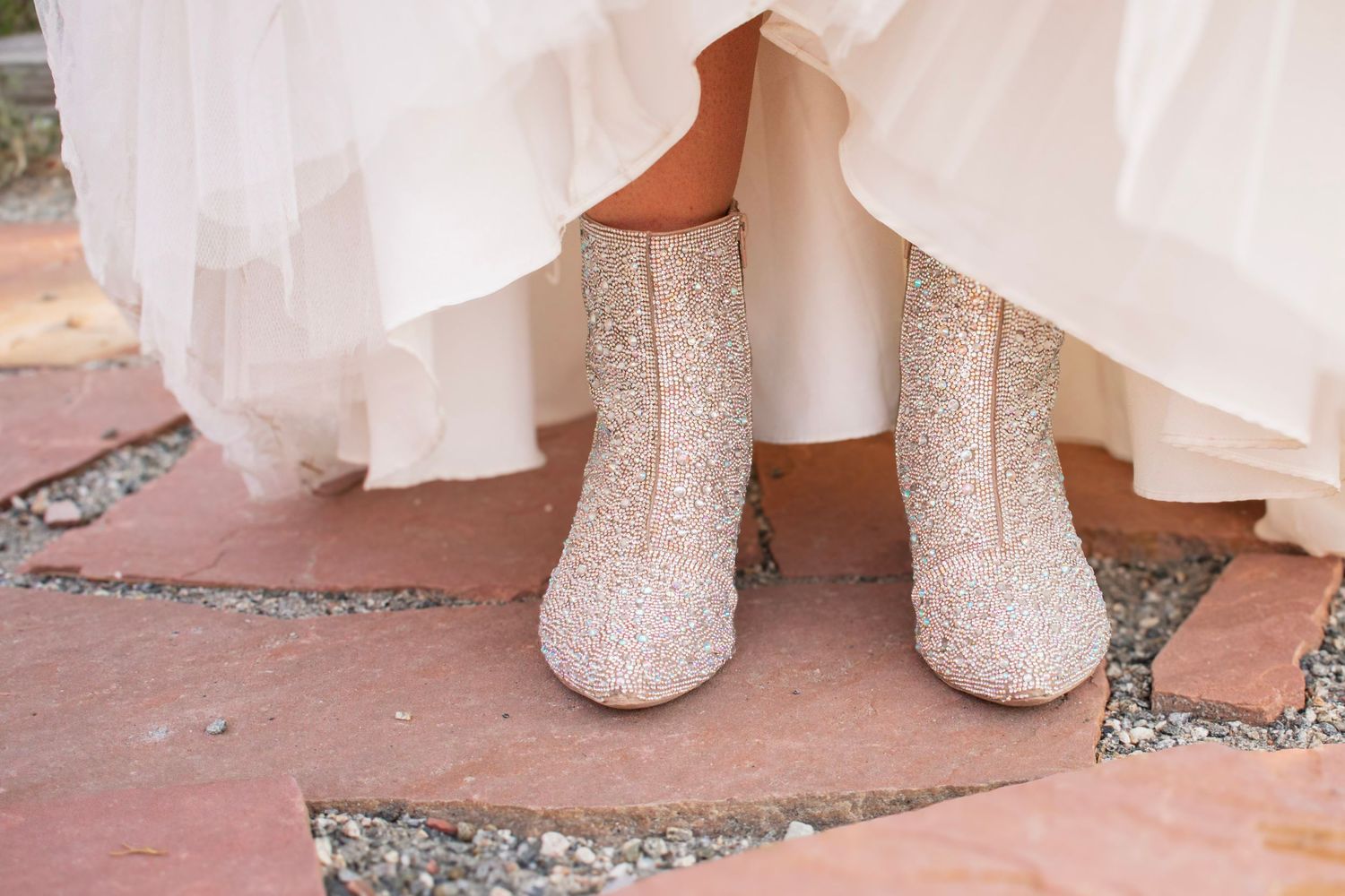 Bride wearing sparkling glitter ankle boots under a white wedding gown on a brick stone pathway.