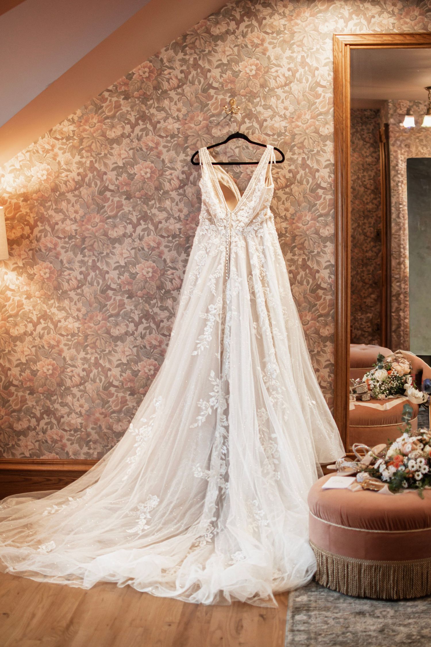 Elegant white lace wedding gown with deep V-neck and flowing train hanging in a romantic floral-wallpapered bridal suite.