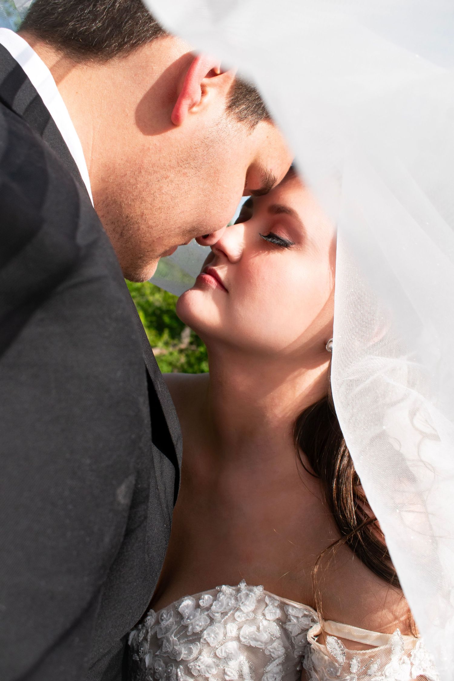 Bride and groom share a tender kiss under a flowing white veil in bright sunlight outdoors.