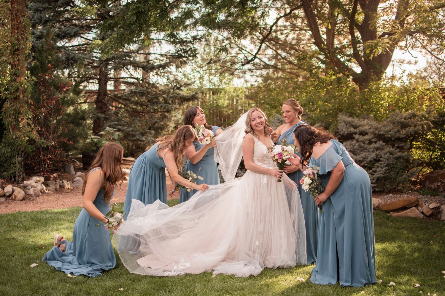 Bride in white gown with bridesmaids in blue dresses arranging veil outdoors on green lawn with trees.