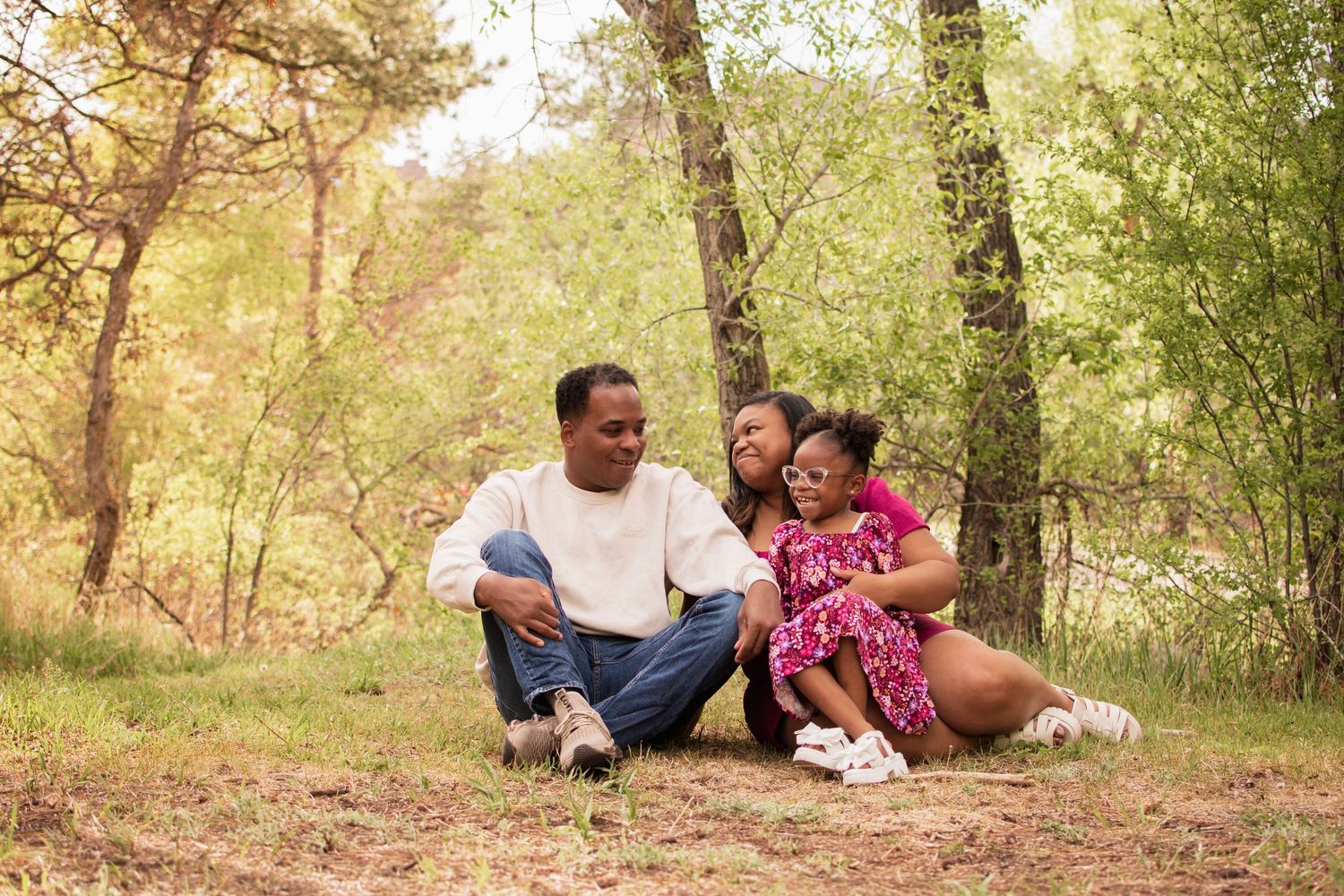 A happy family of three sits together outdoors in a lush forest setting, smiling and embracing on a warm day.