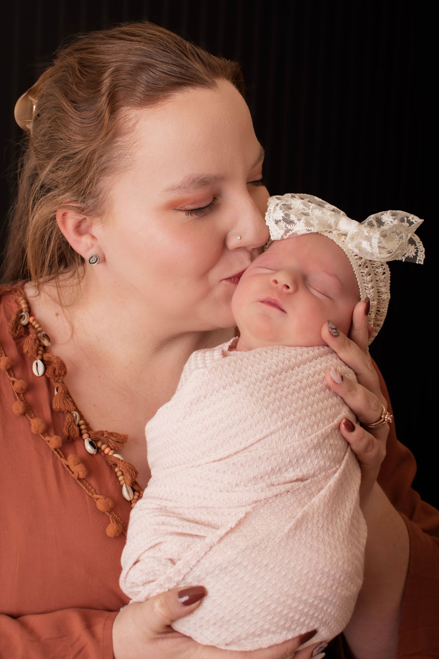 A mother tenderly kisses her swaddled newborn baby wearing a lace bow headband against a dark background.