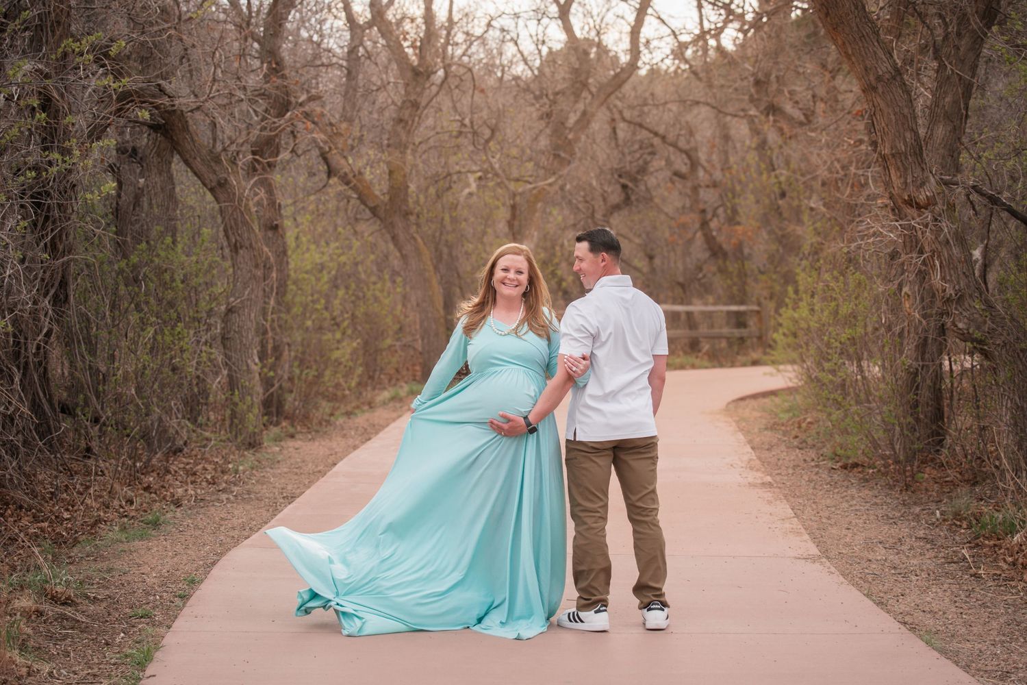 Expecting couple shares a maternity photo moment on a wooded park path, the mother in a flowing mint gown cradling her baby bump.