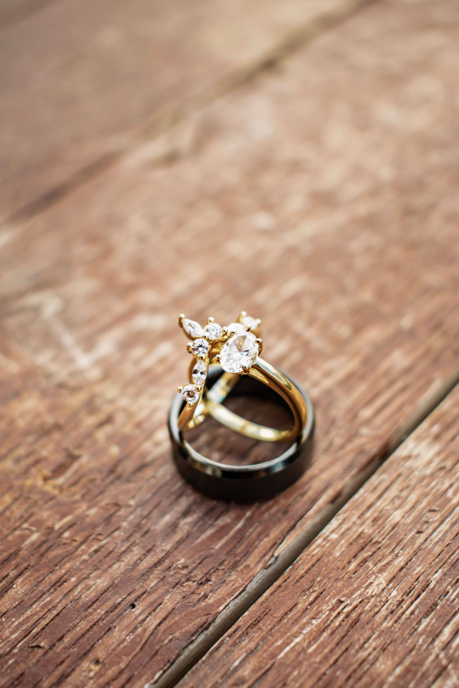 Gold diamond engagement ring and black wedding band resting on rustic wooden surface.