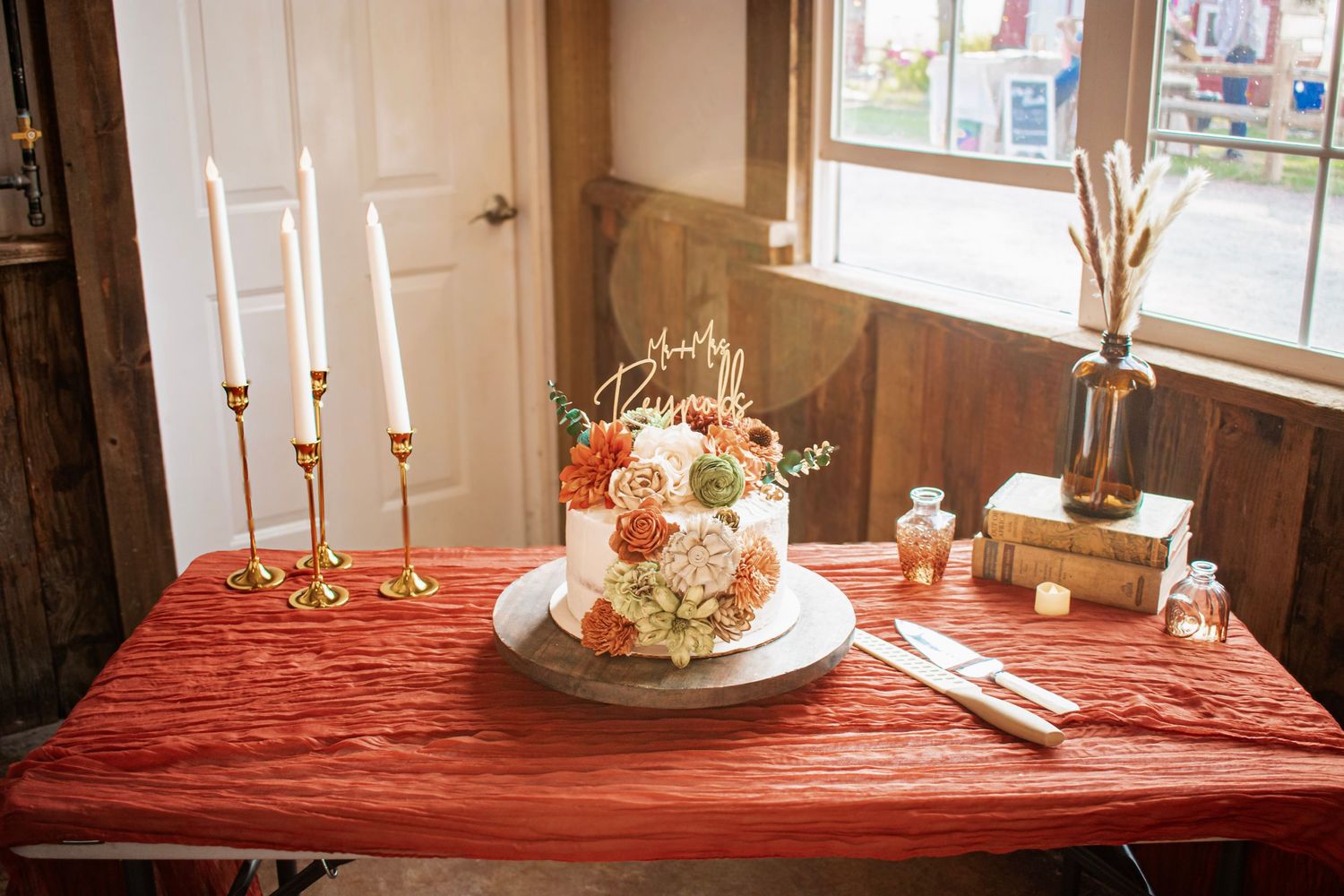 Elegant wedding cake with peach and white florals on a red tablecloth, flanked by gold candlesticks near a sunlit window.