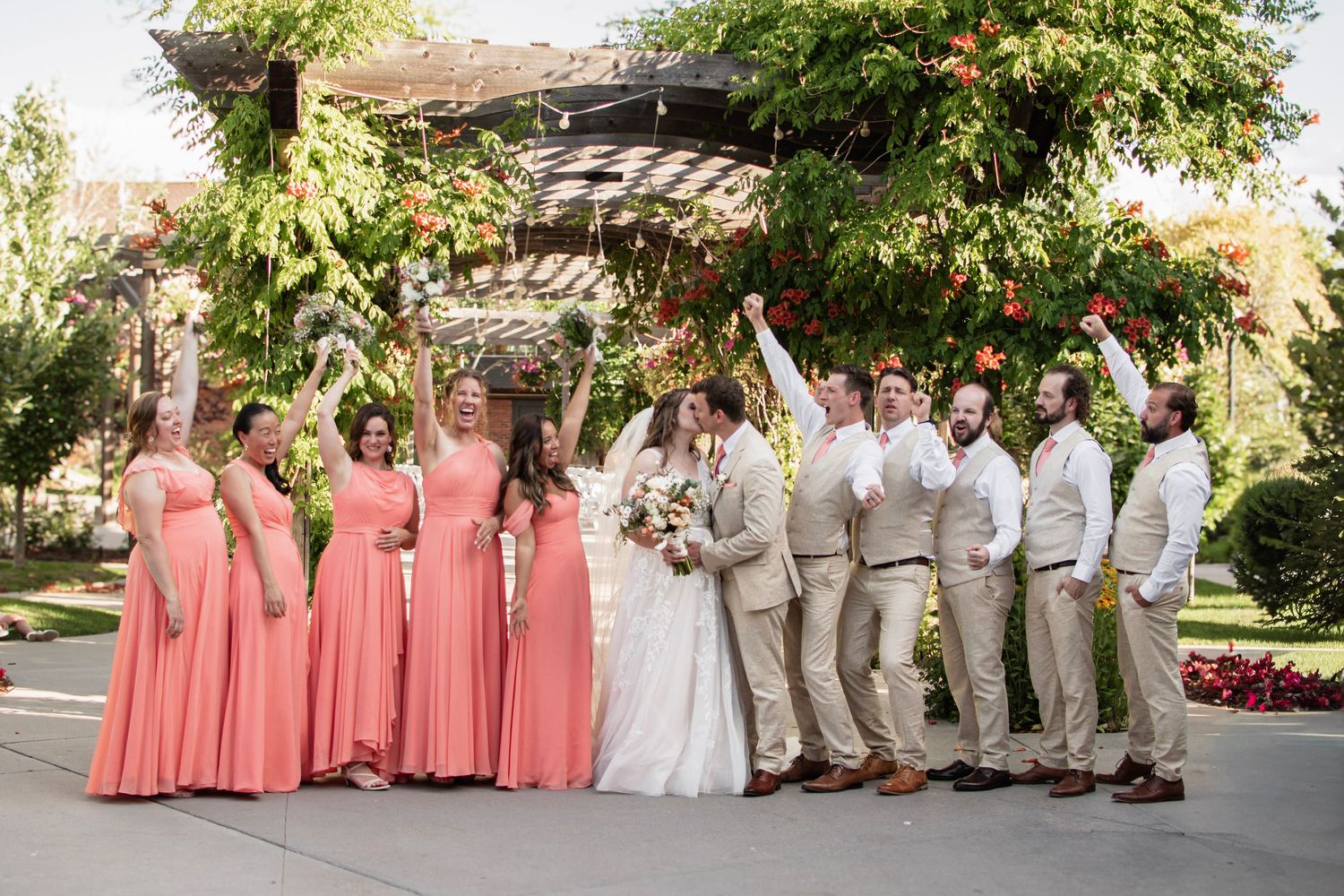 Wedding party celebrates outdoors under floral pergola, bridesmaids in coral gowns and groomsmen in tan suits cheering.