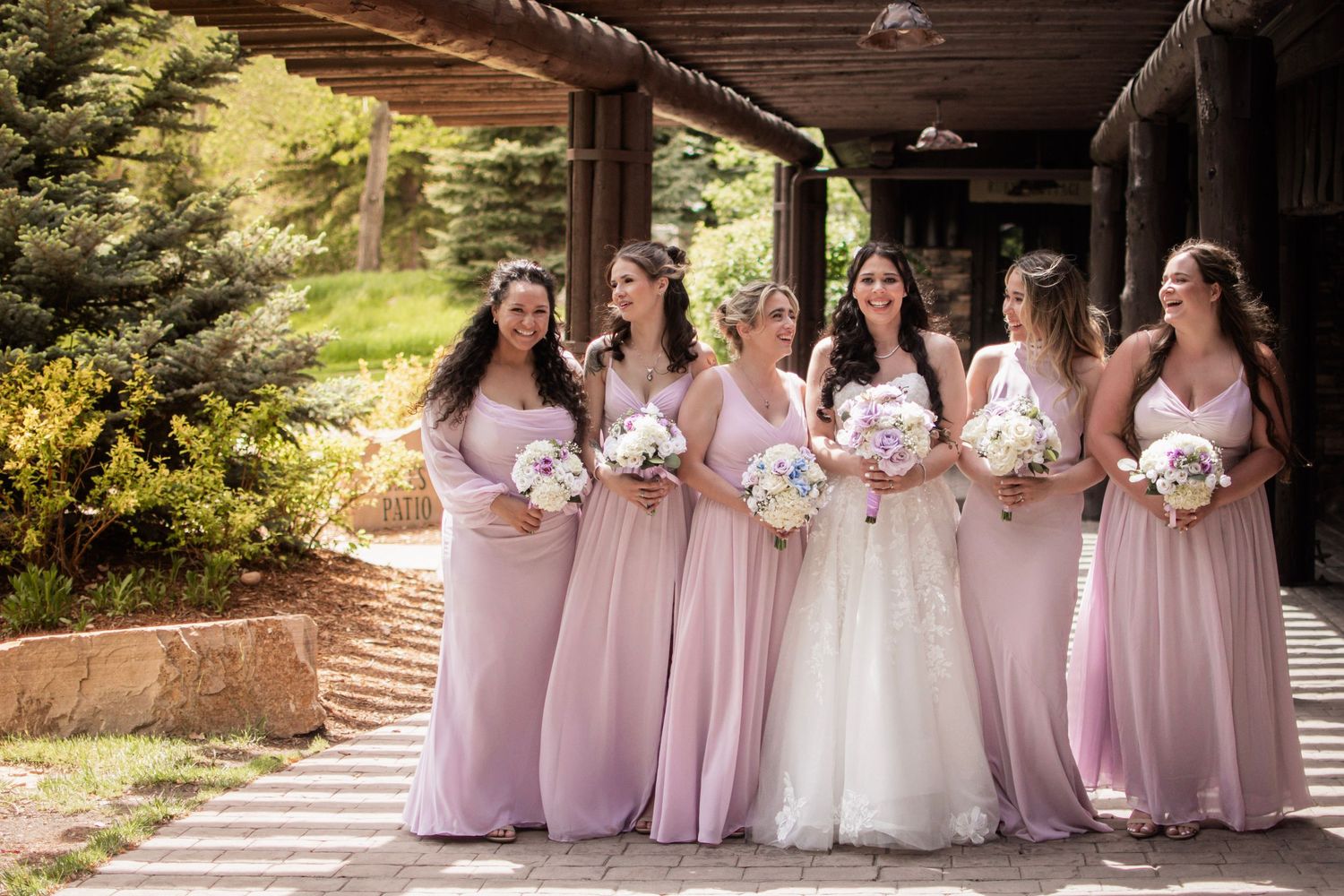 Bride in white gown with five bridesmaids in pink dresses holding bouquets, posing outdoors at a rustic wedding venue.