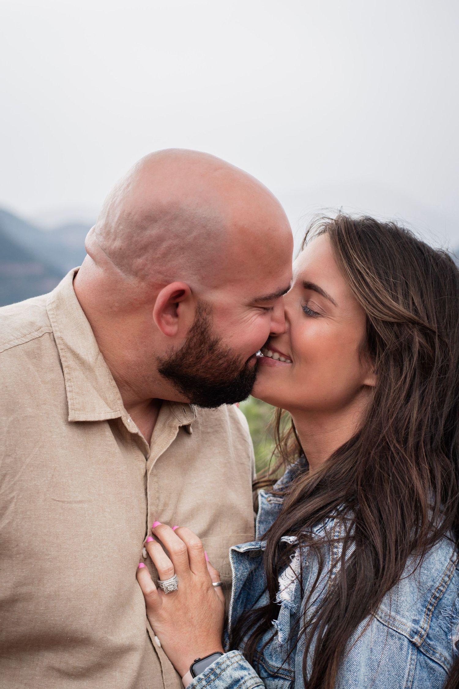 A couple shares a romantic almost-kiss outdoors, woman wearing an engagement ring, mountains visible in the background.