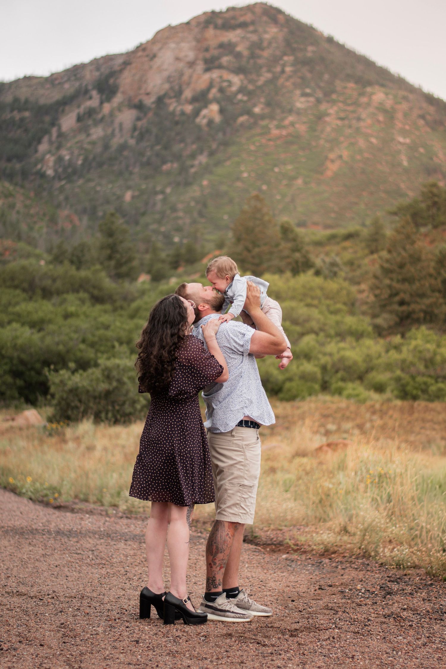 A couple kisses while holding their baby outdoors with a mountain and green trees in the background.