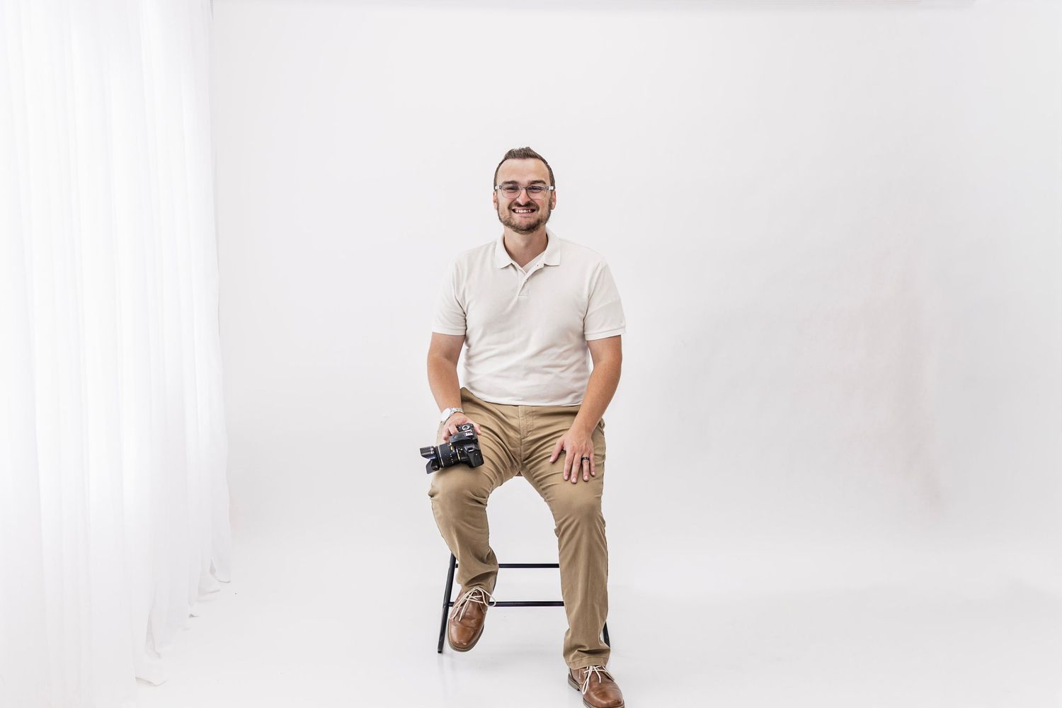 Smiling man in white polo shirt and khaki pants sitting on a stool holding a camera in a bright white studio.