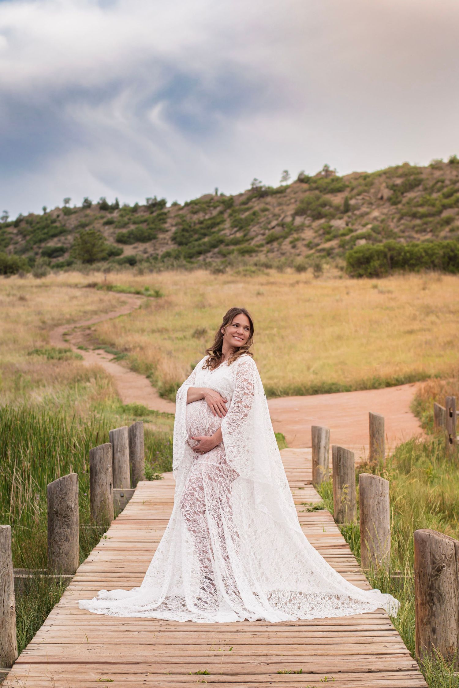 Pregnant woman in white lace gown on wooden bridge, scenic Colorado landscape with hills and cloudy sky at Ute Valley Park.