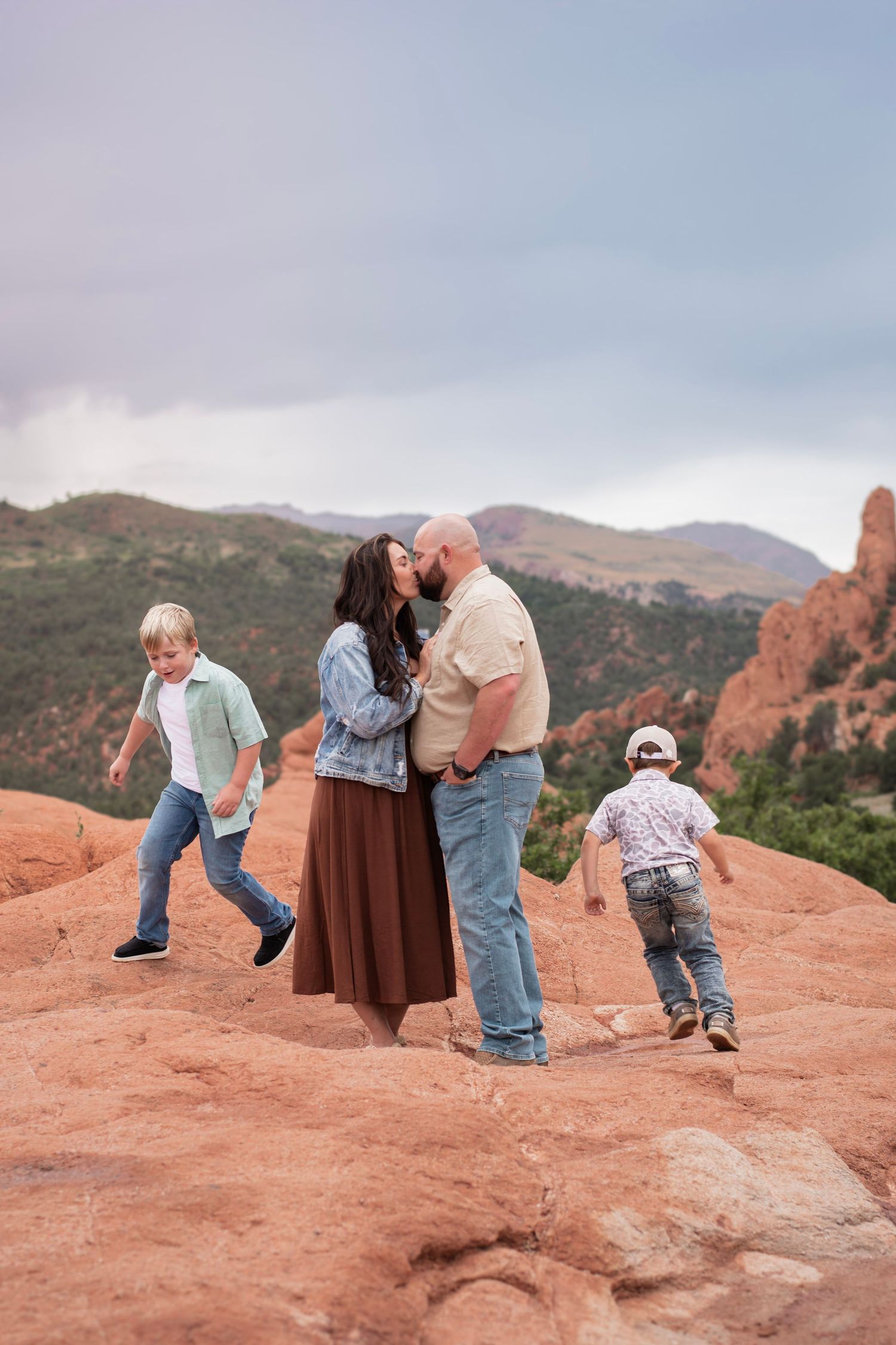 Couple kissing on red rocks at Garden of the Gods while two young boys play around them, mountains in background.