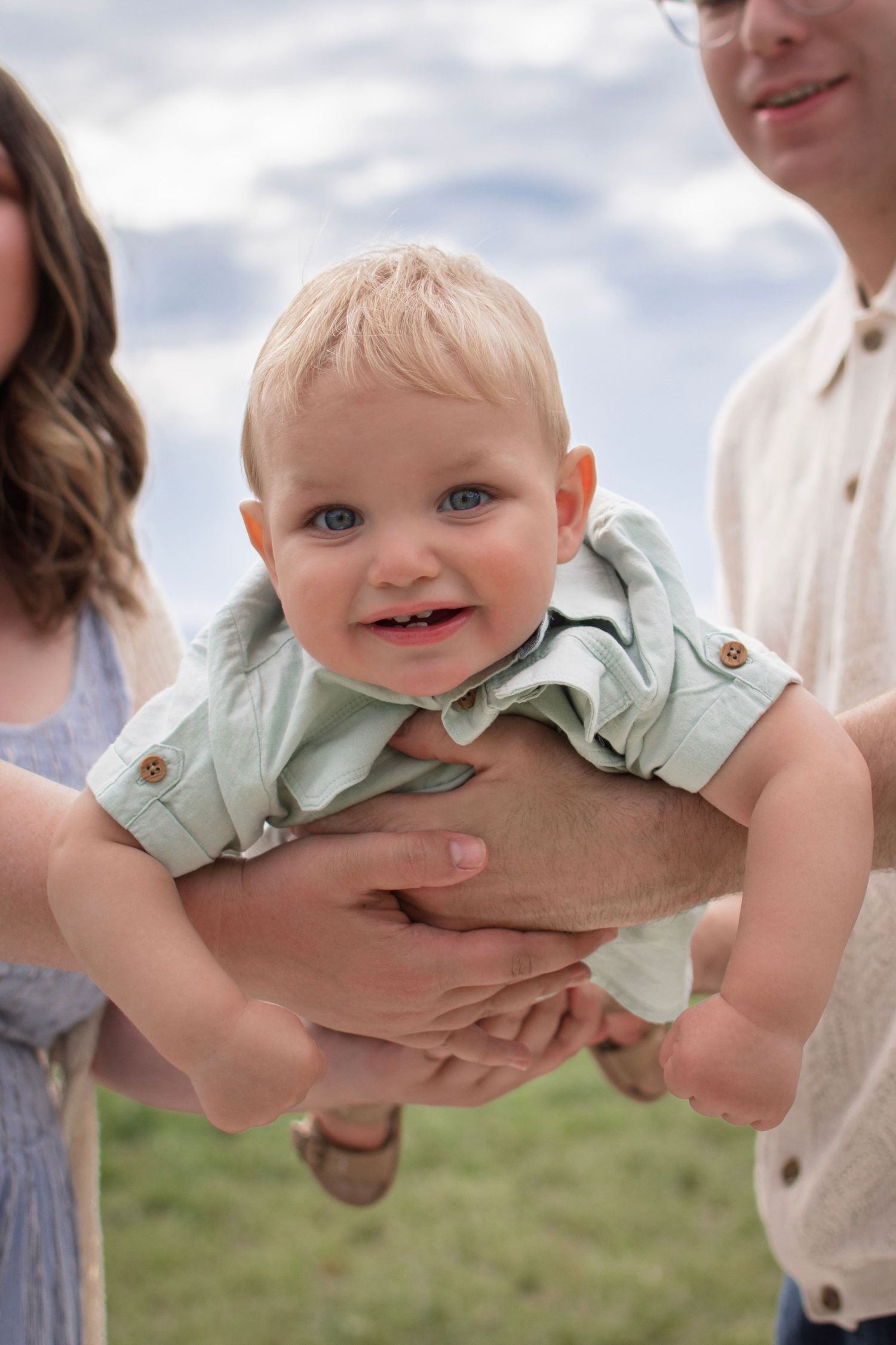 A smiling baby in a green outfit being held up by parents outdoors under a cloudy sky.