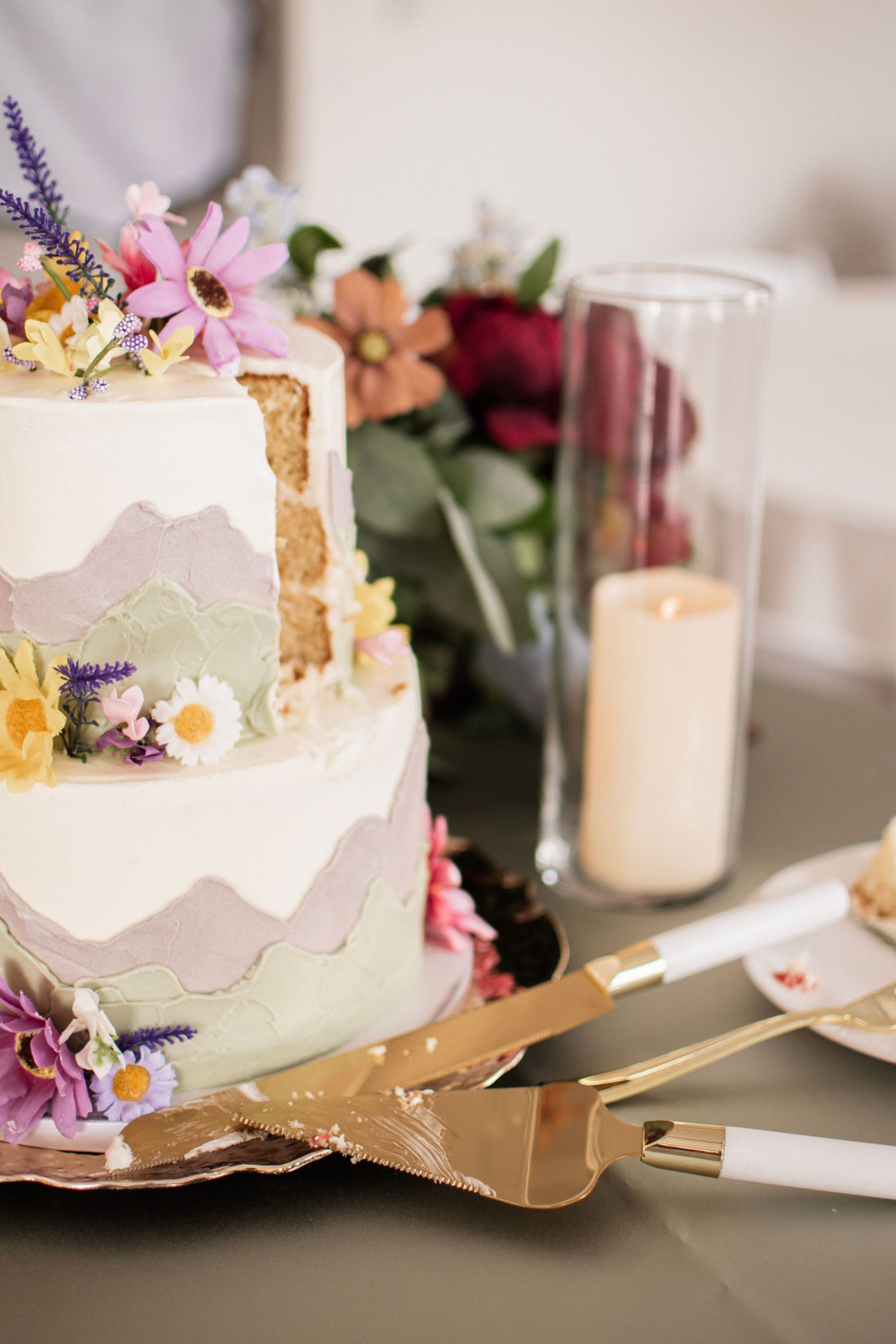 Elegant two-tier wedding cake decorated with colorful wildflowers in purple, pink, and yellow with gold serving set.