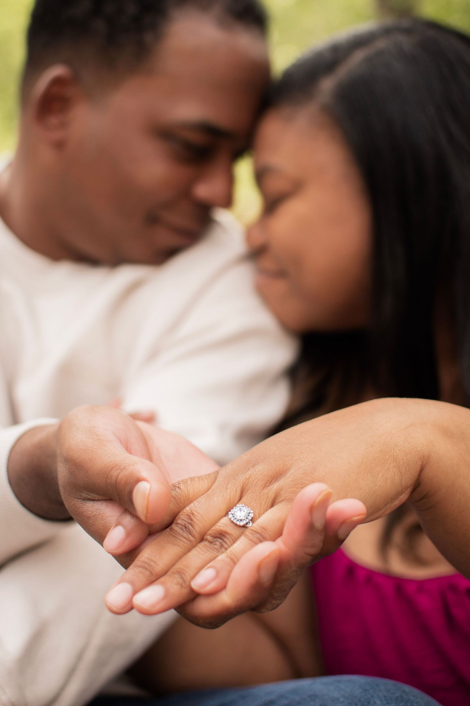 Couple holding hands showing engagement ring while leaning foreheads together outdoors.