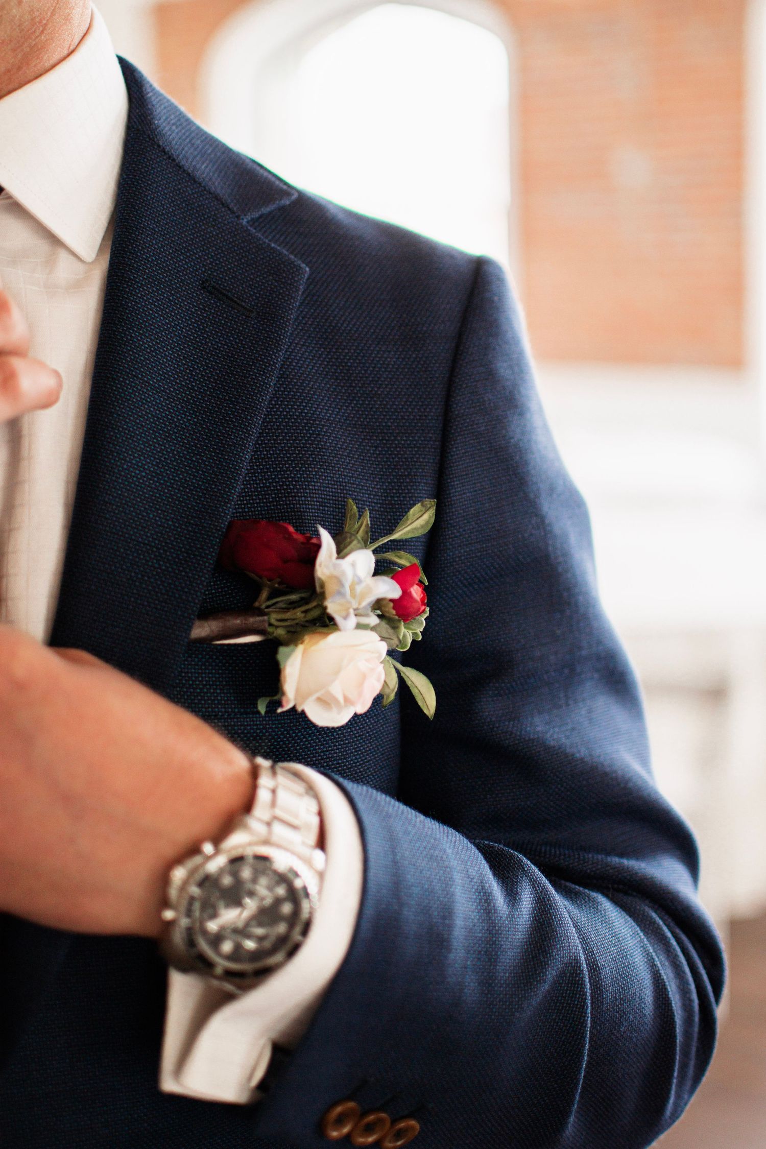 Groom in navy blue suit with red and white floral boutonniere, wearing a silver watch, adjusting his jacket lapel.