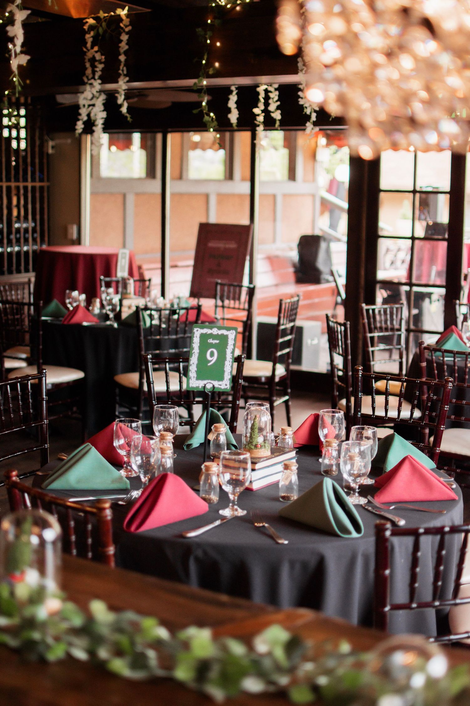 Elegant wedding reception venue with black tablecloths, red and green napkins, chiavari chairs, and string lights overhead.