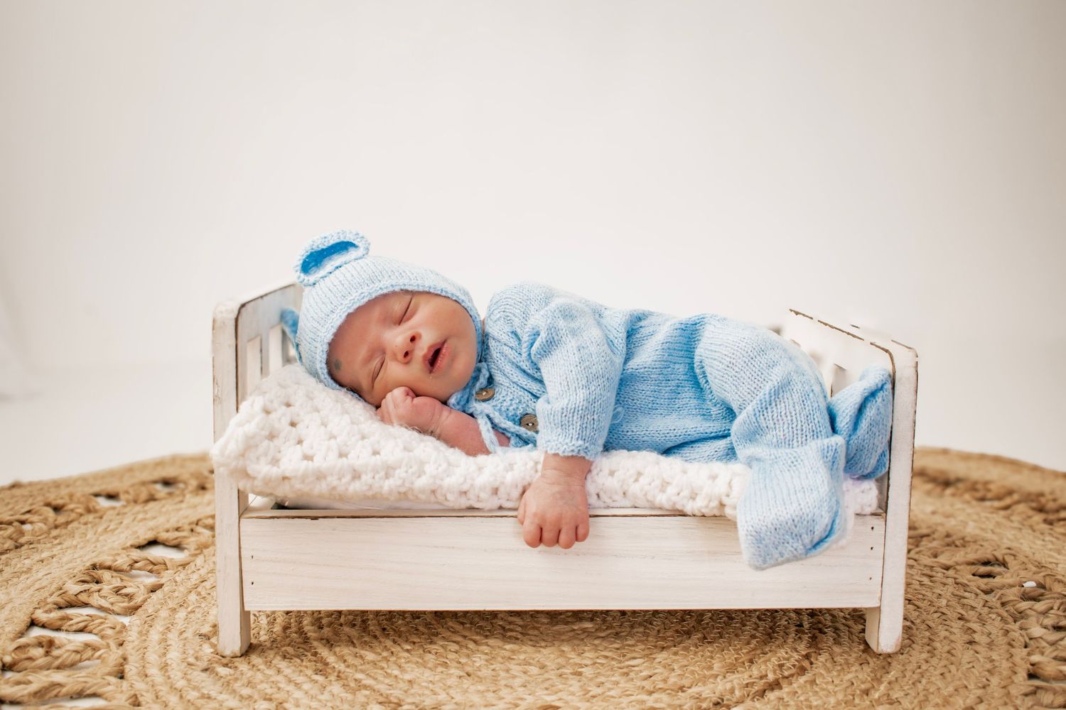 Newborn baby sleeping peacefully in blue knit outfit on a tiny white wooden bed with a woven rug.