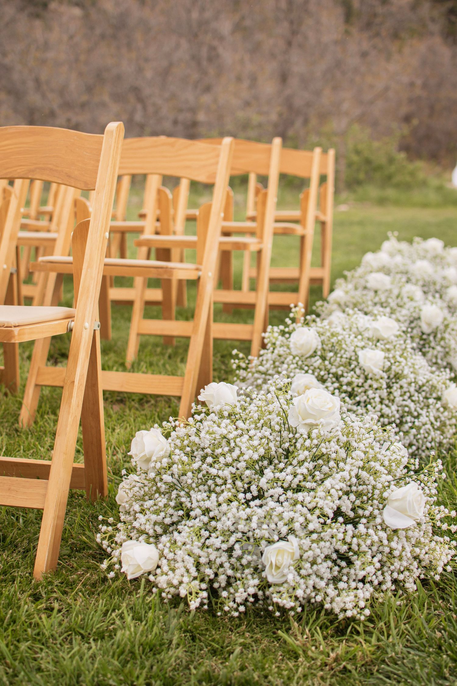 White roses and baby's breath floral arrangement lining outdoor wedding ceremony aisle with wooden chairs.