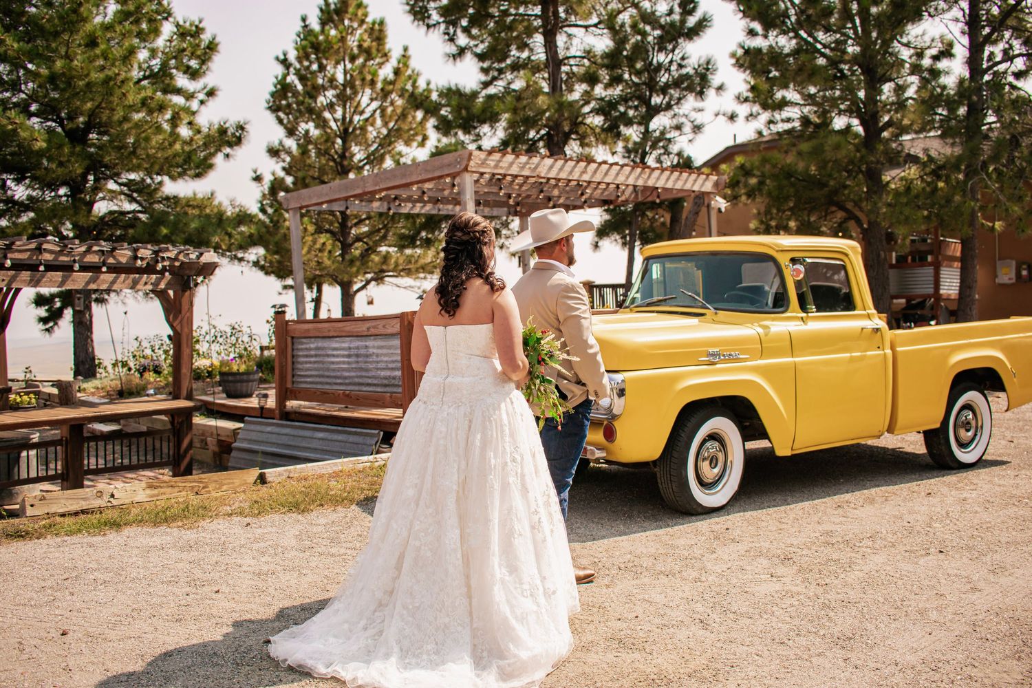Bride in white gown and groom in cowboy hat walk toward a vintage yellow pickup truck at outdoor wedding venue.