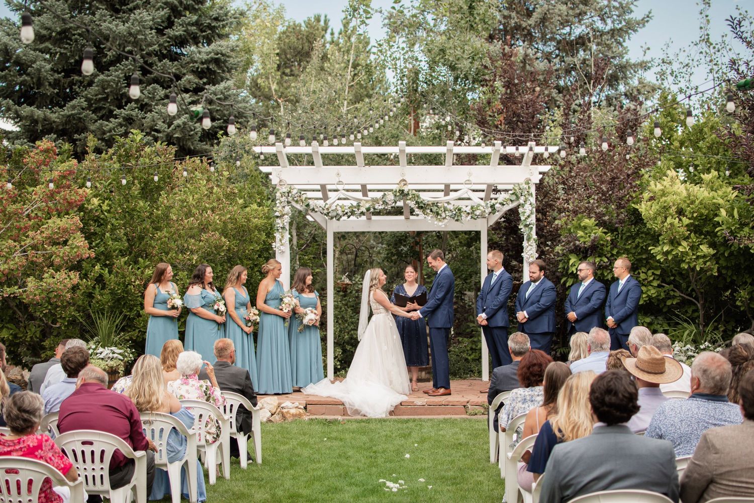 Outdoor wedding ceremony under white pergola with bridesmaids in blue dresses and groomsmen in navy suits surrounded by lush greenery.