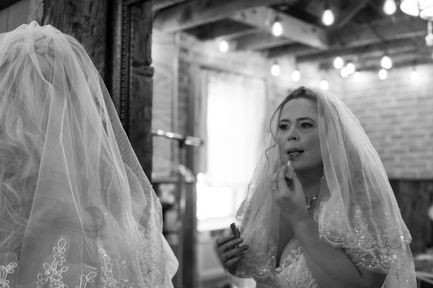 Black and white portrait of bride in lace gown and veil touching up lipstick while looking in mirror.