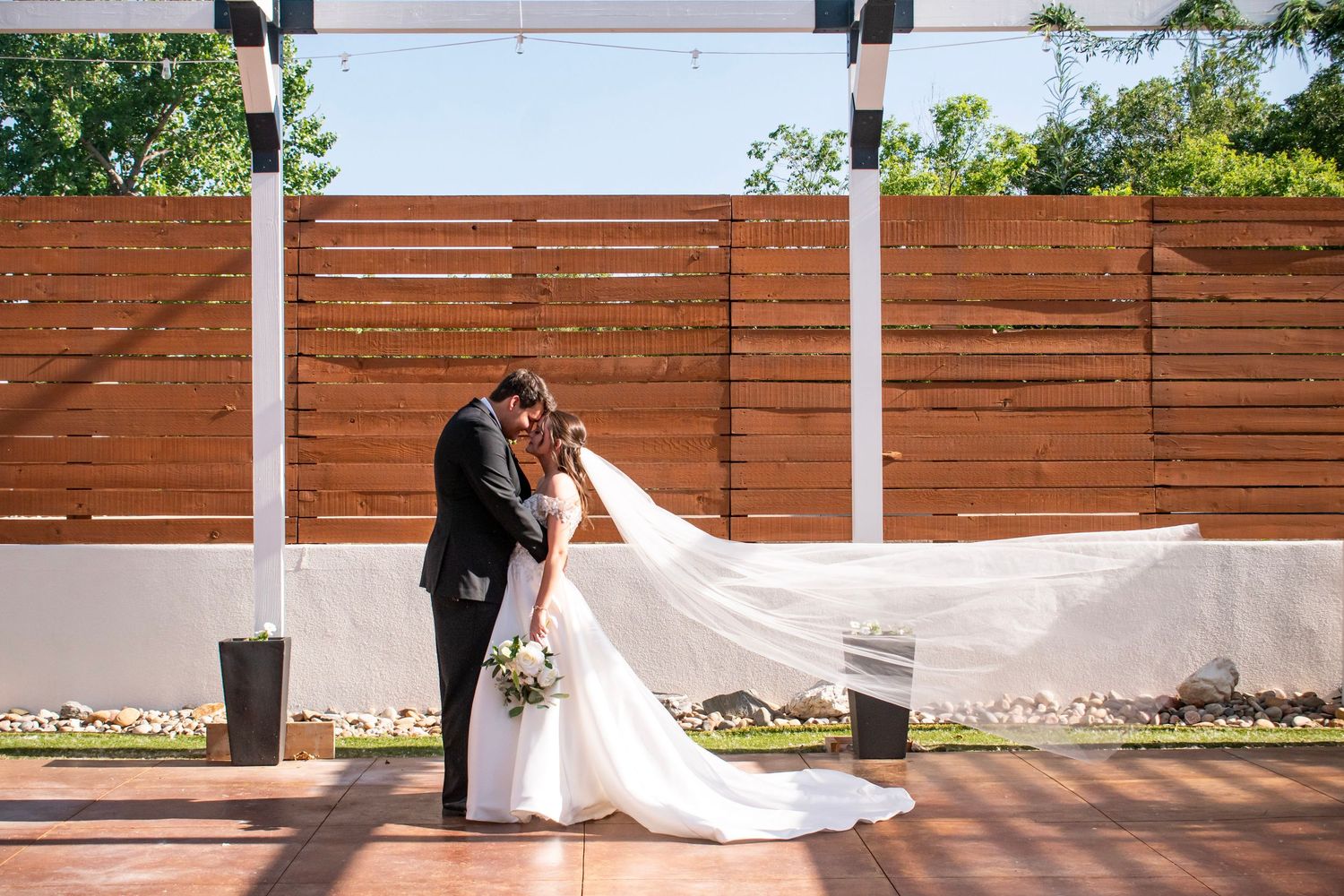 Bride and groom share a romantic kiss outdoors, with the wedding veil flowing in the wind against a wooden fence backdrop at Creekside