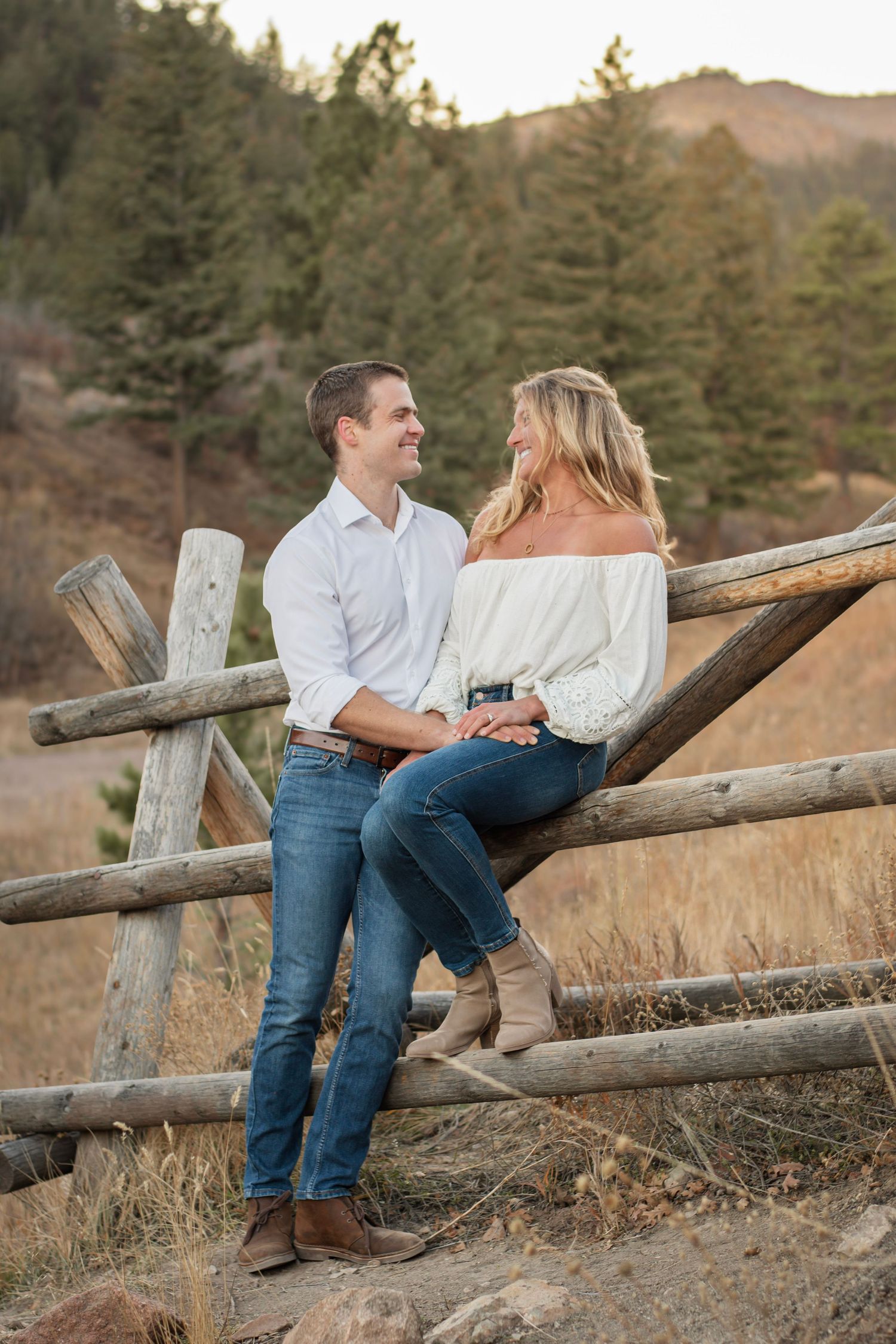 A couple smiles at each other while sitting on a rustic log fence in a scenic mountain meadow setting.