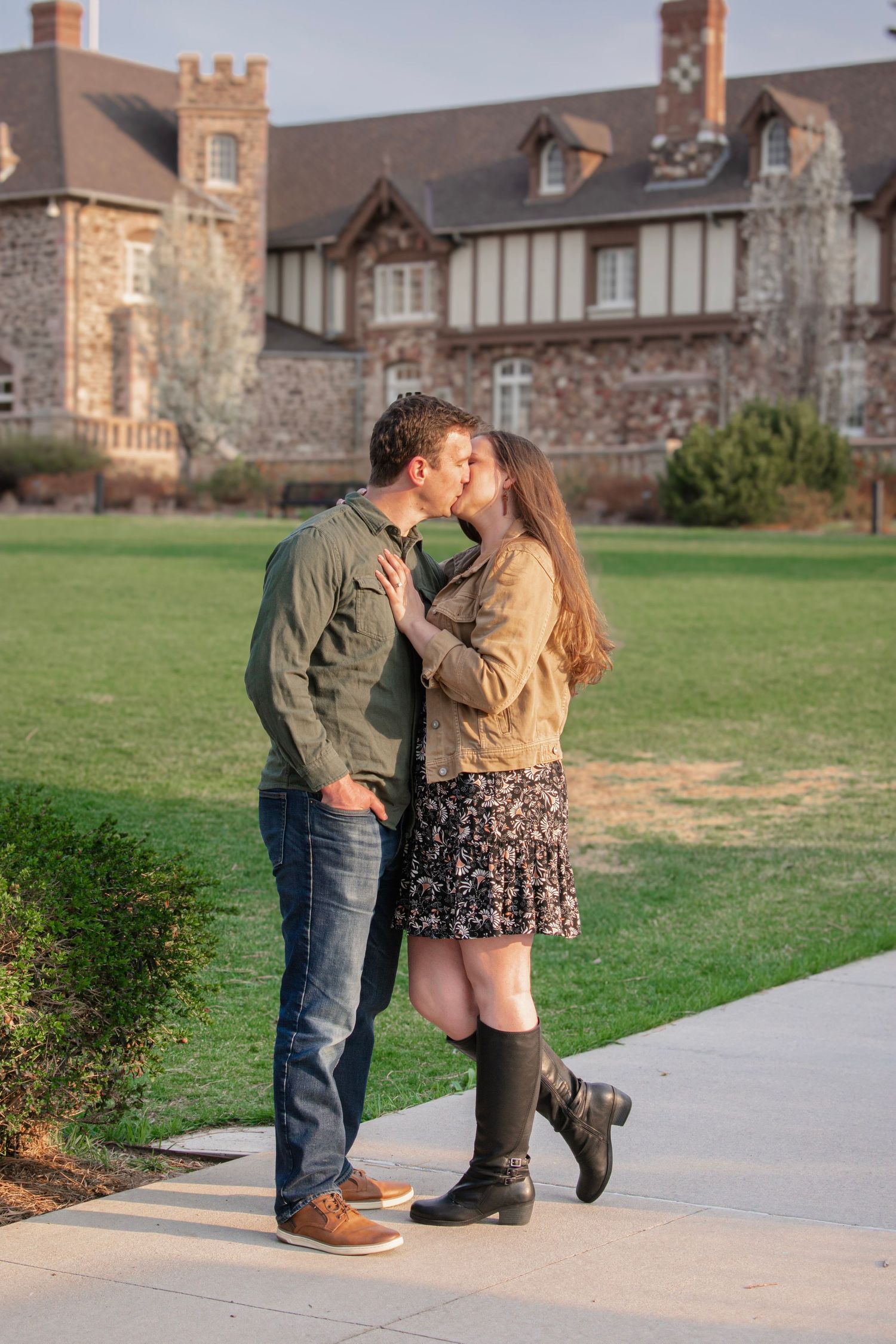 Couple sharing a romantic kiss on a pathway outside a grand stone Tudor-style building with lush green lawn, at Highlands Ranch Mansion.