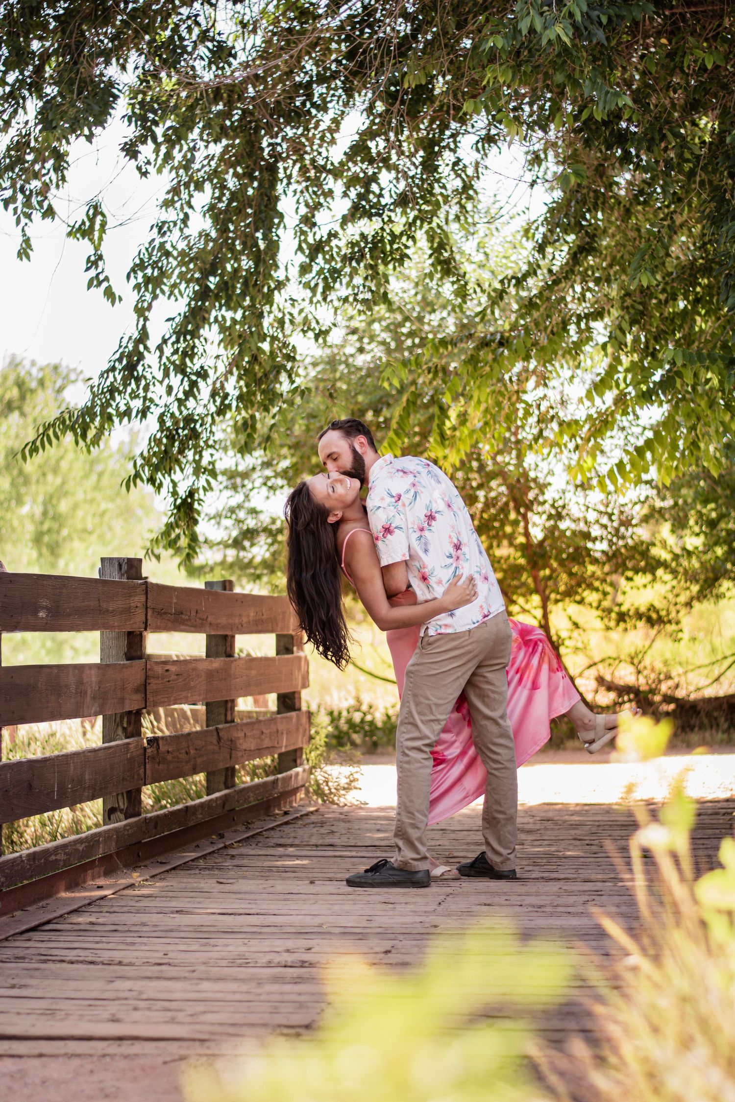 Couple sharing a romantic kiss on a wooden bridge surrounded by lush green trees in a sunny outdoor setting.