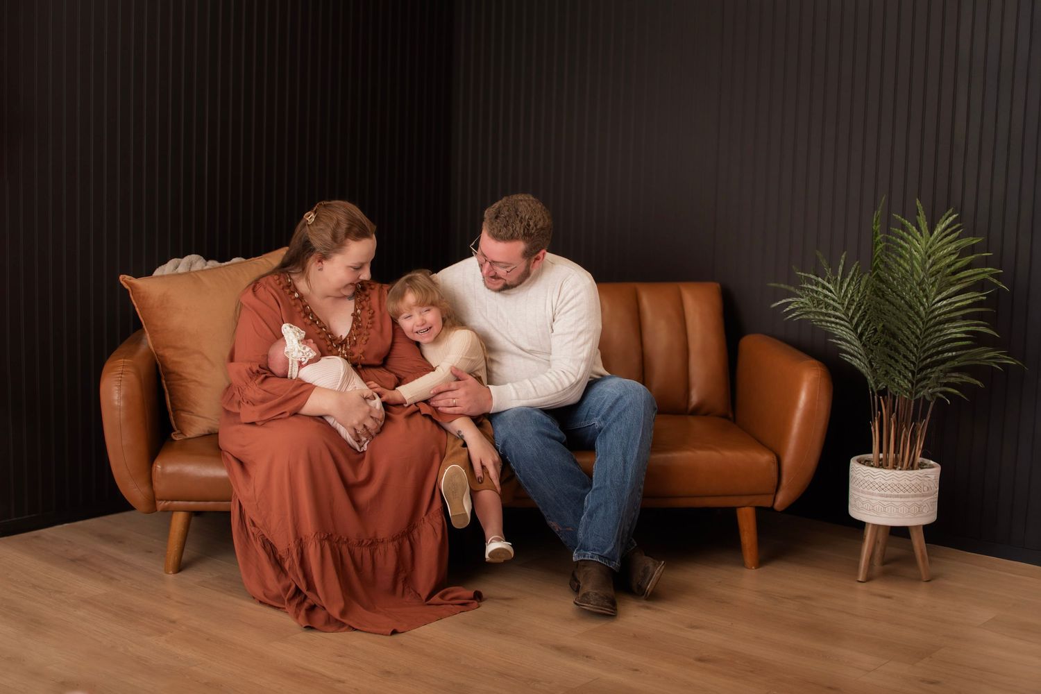 Family of three sitting on brown leather sofa, parents admiring newborn baby, toddler beside them, against dark wall.