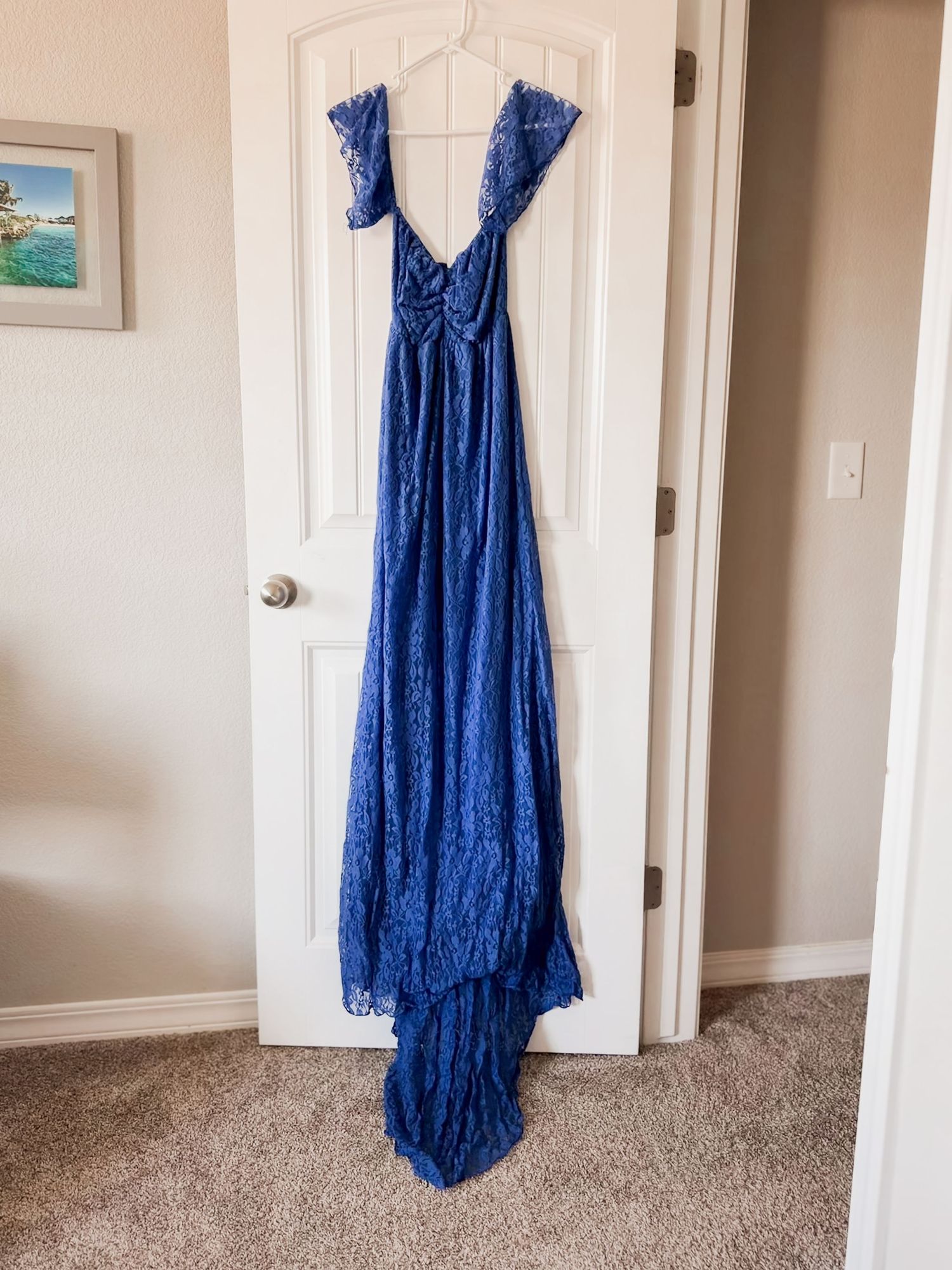 Royal blue lace maxi dress with cap sleeves and a flowing train hangs on a white paneled door in a bedroom with beige carpet.