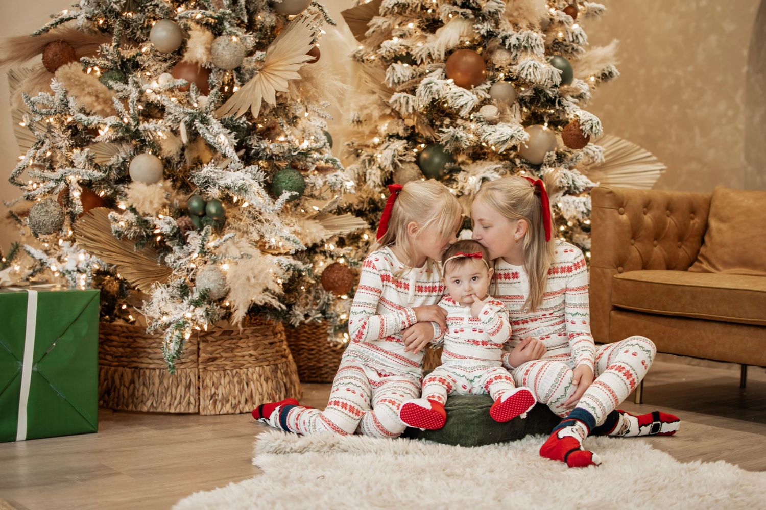 Three children in matching Christmas pajamas sit together by a decorated holiday tree surrounded by wrapped gifts.