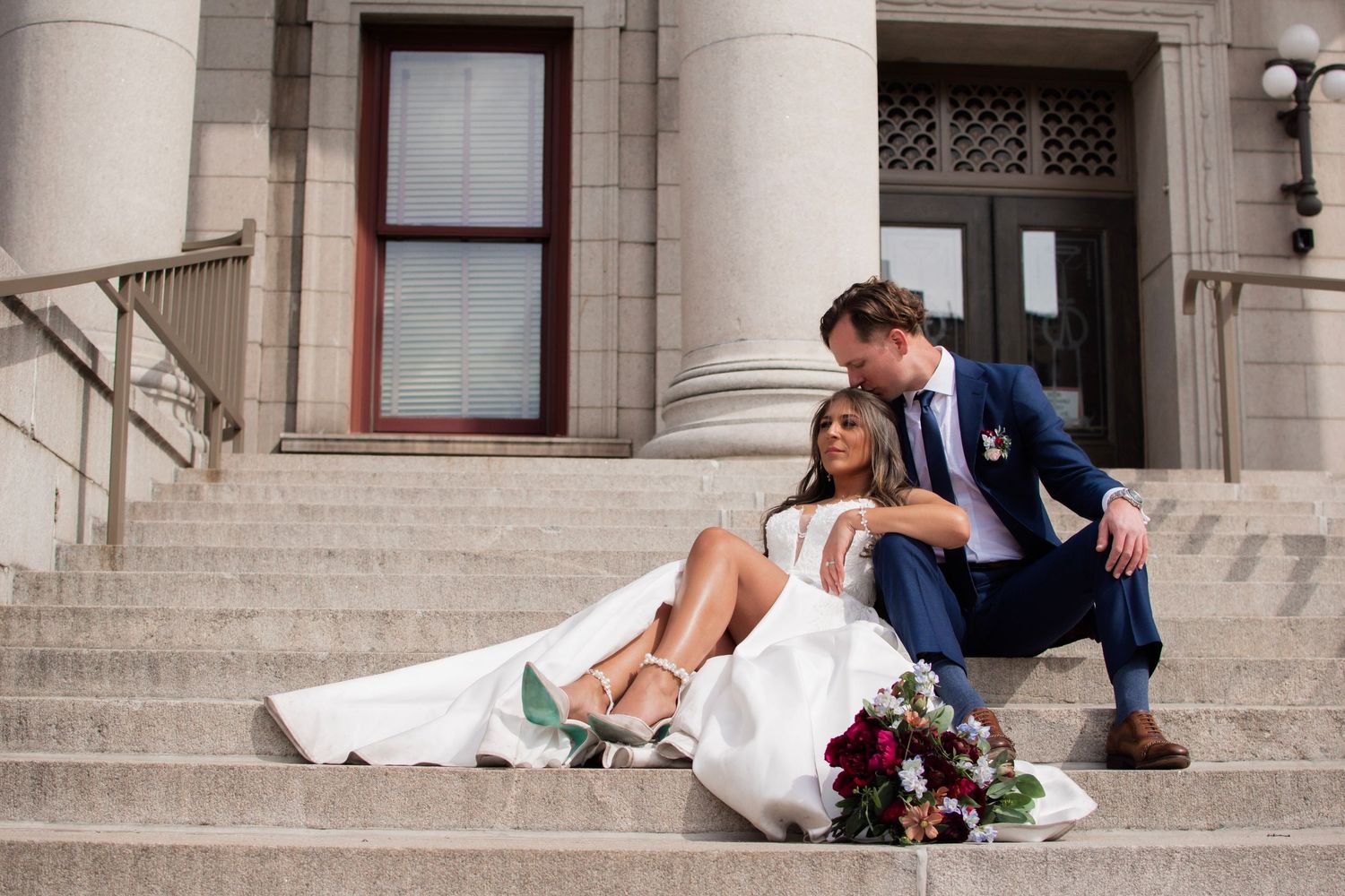 Bride and groom sit together on stone steps of a grand building with a bouquet of red flowers.