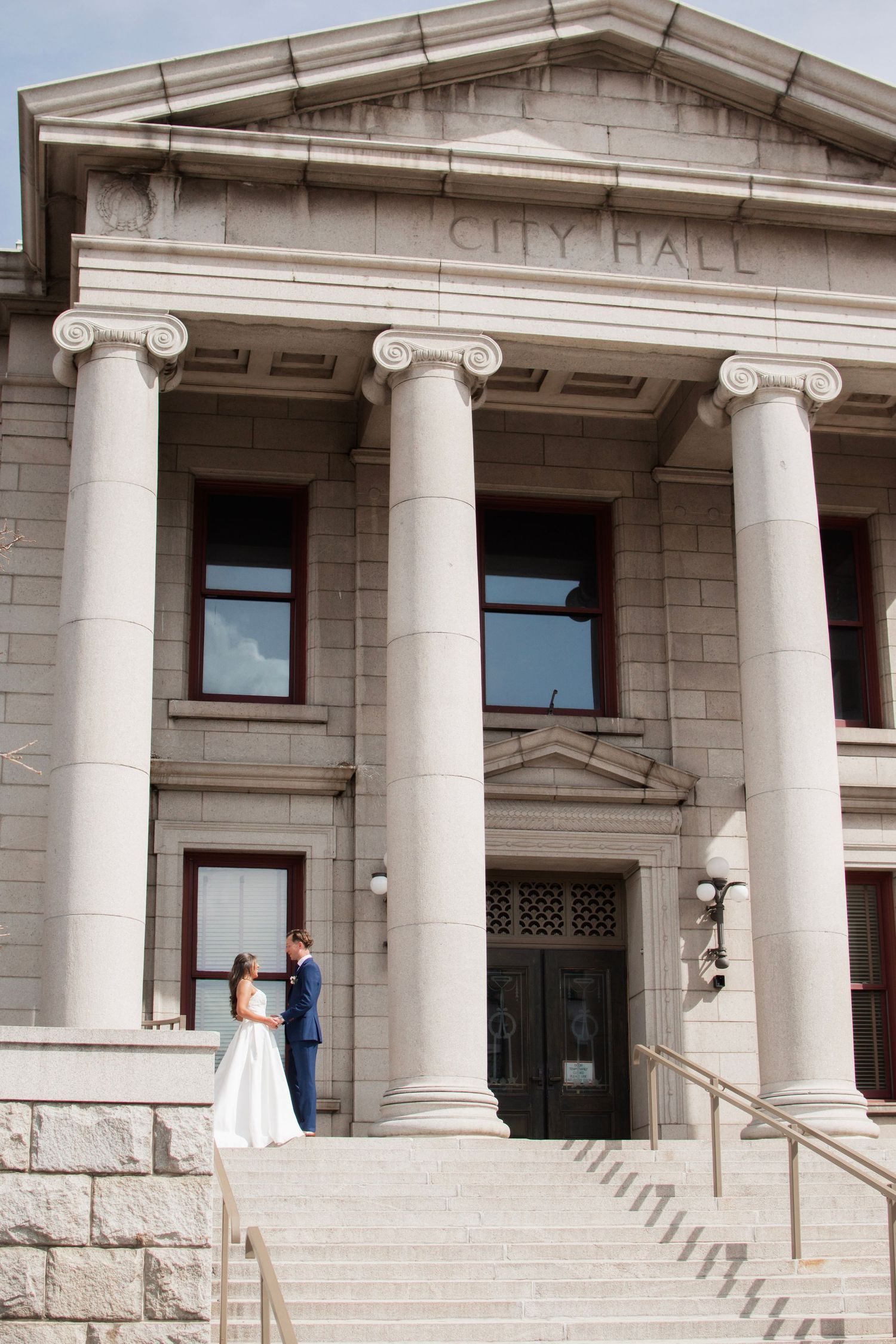 Bride and groom pose on steps of historic City Hall building with grand columns on their wedding day.