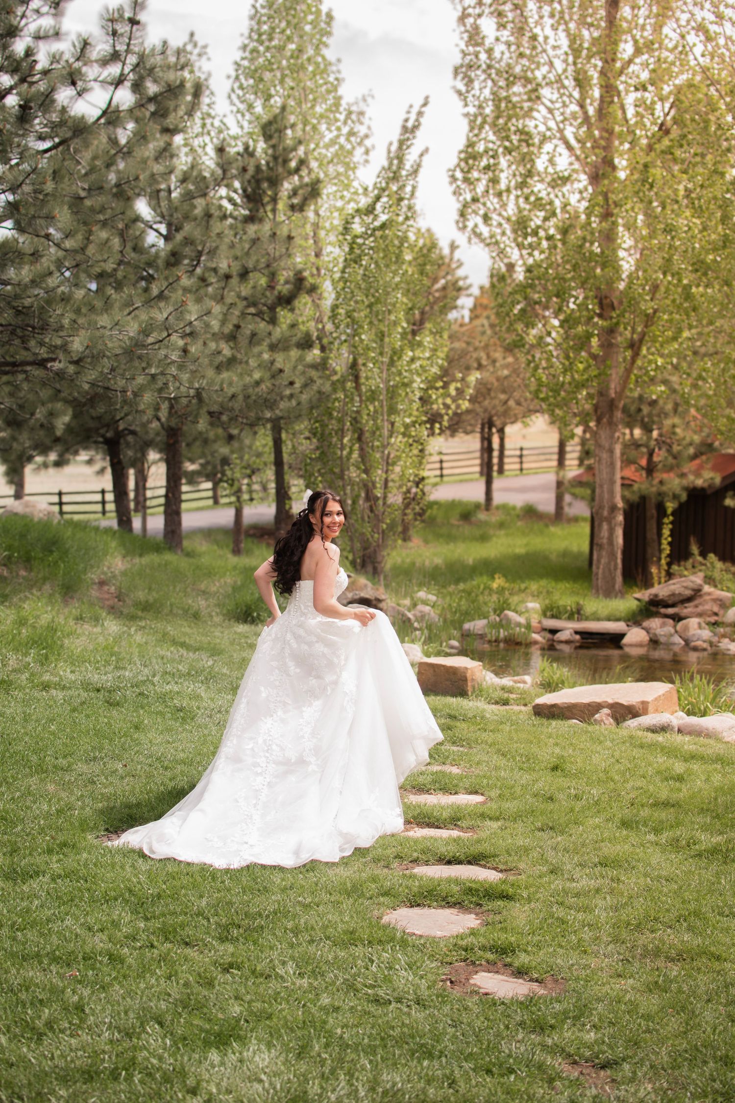 Bride in white ball gown standing on garden stepping stones near a pond surrounded by lush green trees.