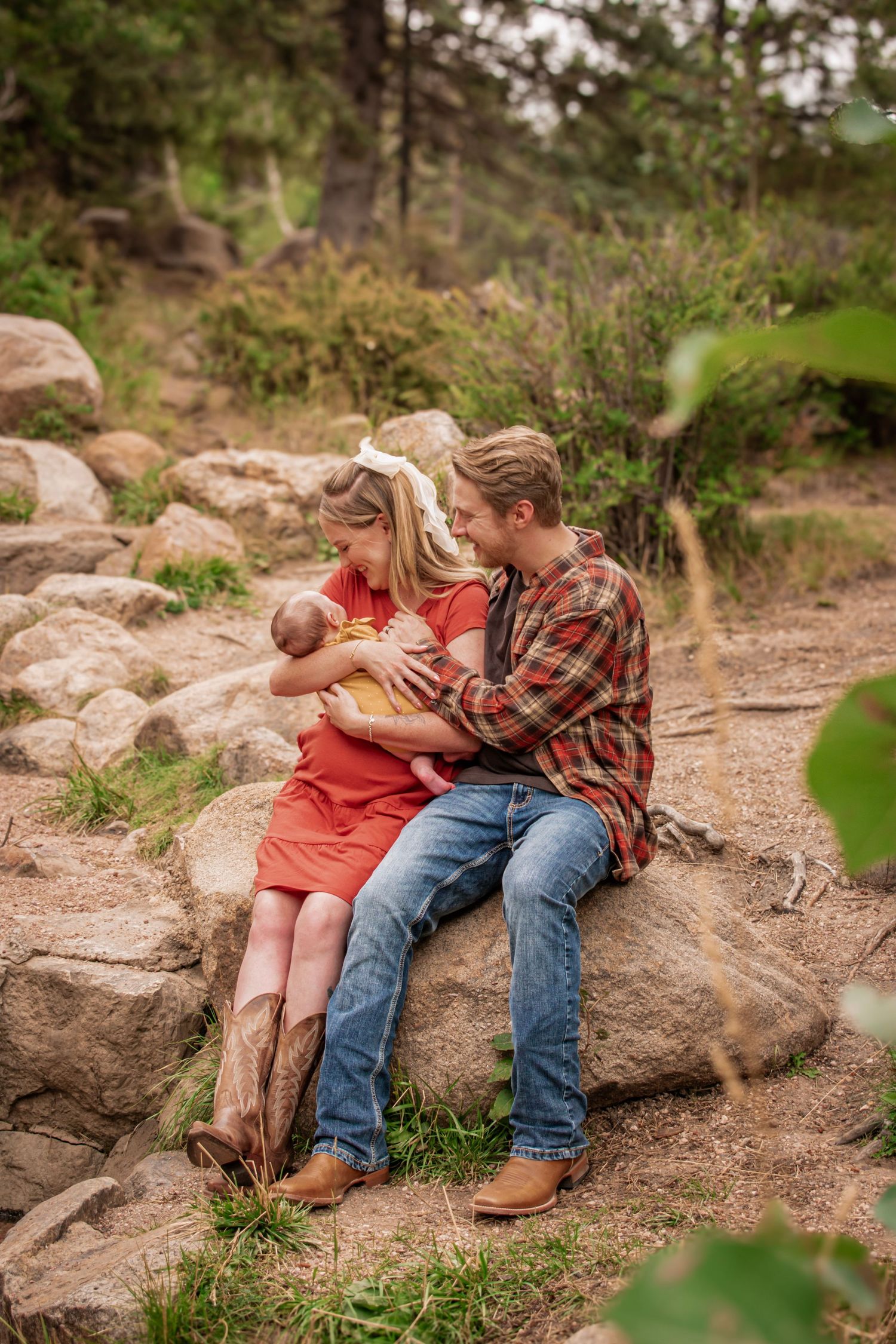Young couple in western attire sitting on rocks in nature, lovingly holding their newborn baby outdoors.