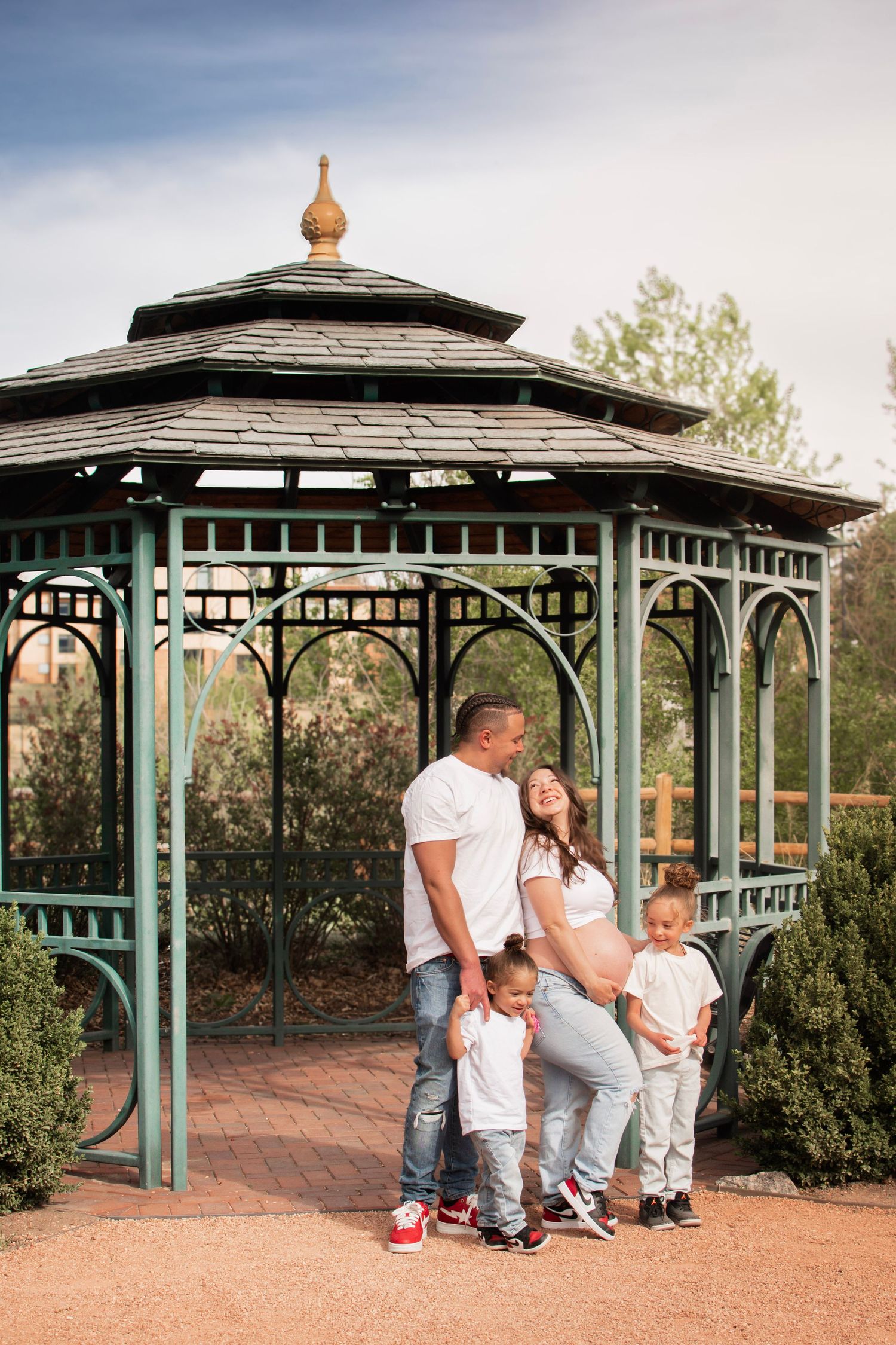 Pregnant woman and family posing in front of a green gazebo in a park.